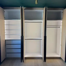 Otwarta szafa wnękowa z oświetleniem LED, widoczne półki, szuflady i drążek na ubrania. Jasne drewno frontów kontrastuje z ciemnozieloną ścianą.