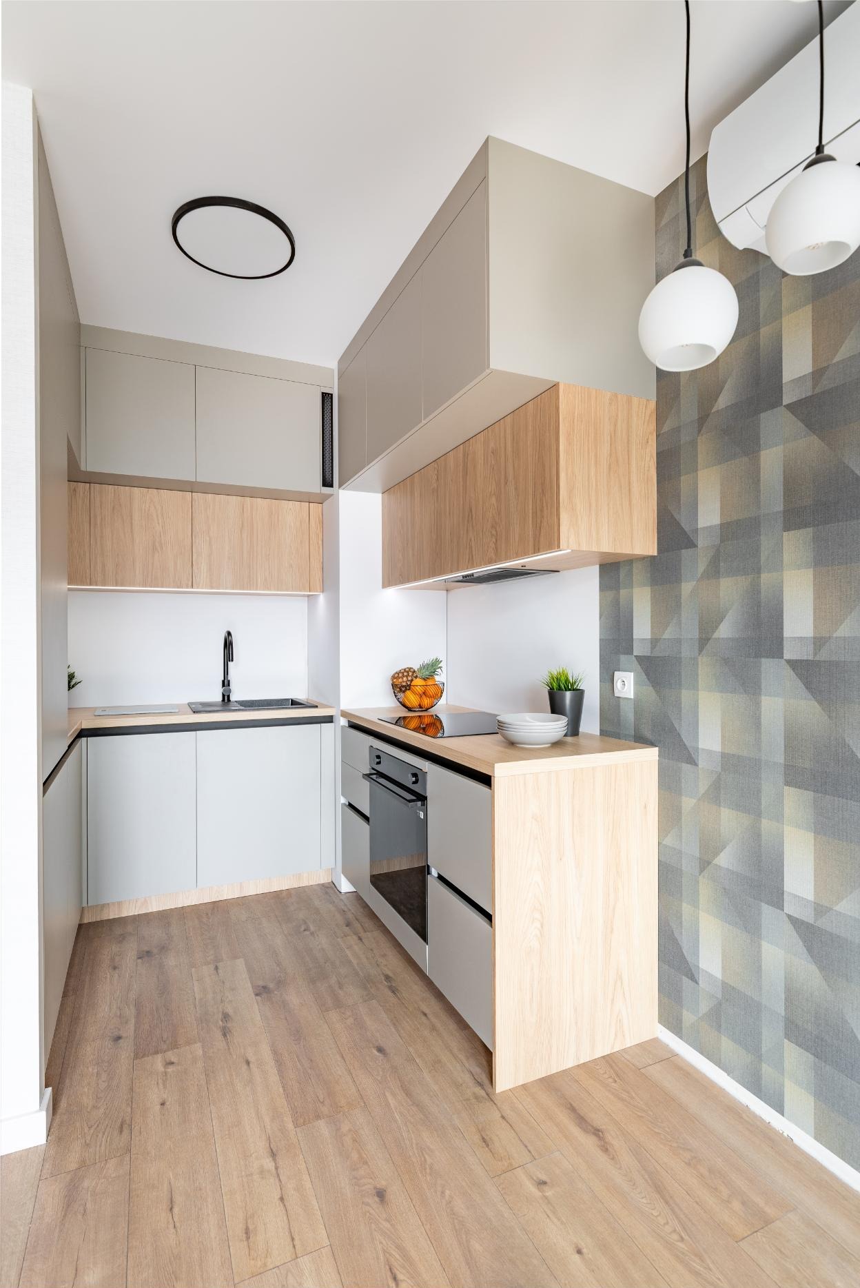 Nowoczesna kuchnia z drewnianymi i szarymi szafkami, czarną armaturą i geometryczną tapetą. Oświetlenie punktowe i drewniana podłoga dodają ciepła wnętrzu.