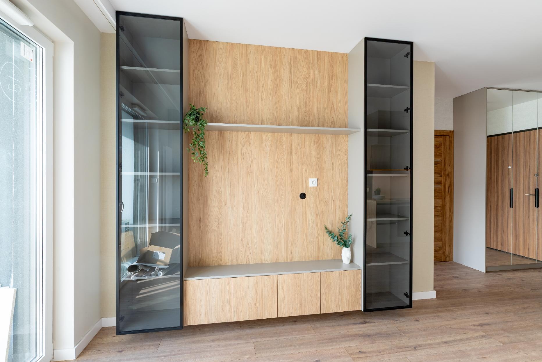 Nowoczesna zabudowa meblowa z drewnianą ścianką, szklanymi witrynami w czarnych ramach i półką. Minimalistyczny design, jasne drewno, roślina doniczkowa. Parkiet na podłodze.