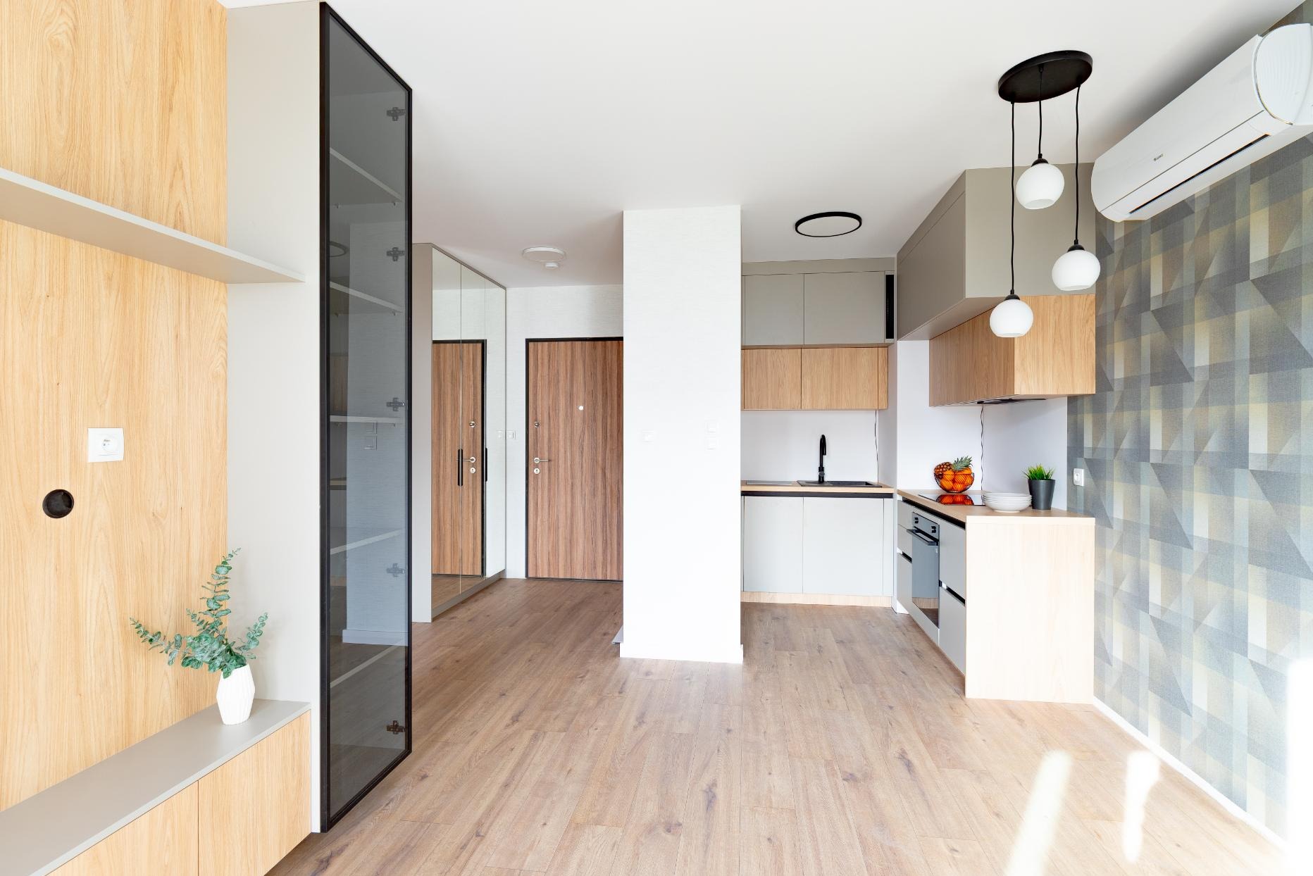 Jasne wnętrze: zabudowa z drewnianym fornirem, szklana witryna, lustro, drzwi wejściowe, kuchnia z szarymi szafkami i drewnianymi akcentami, lampy wiszące, klimatyzator.