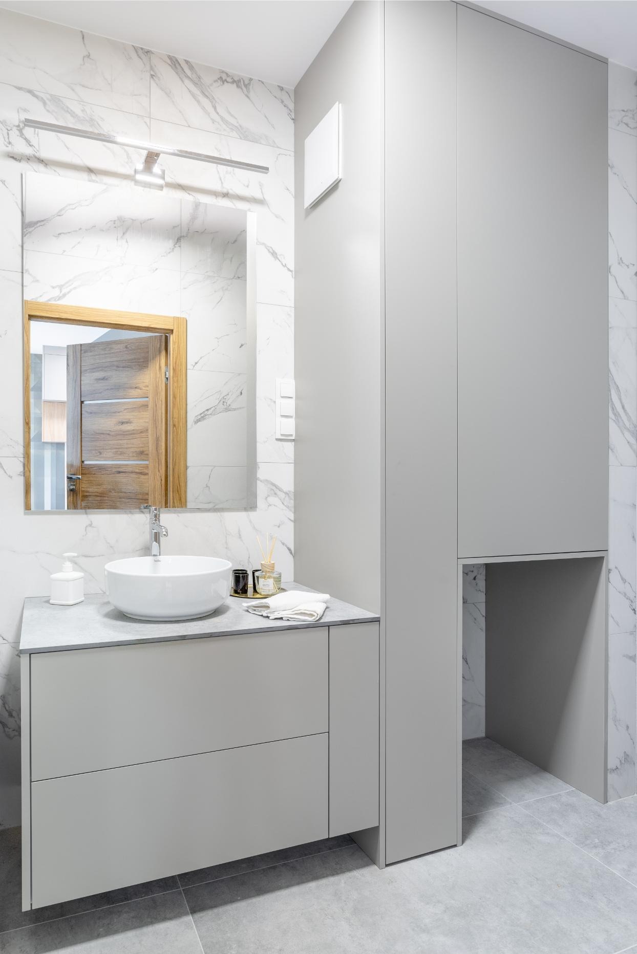 Nowoczesna łazienka w szarościach z marmurową ścianą, okrągłą umywalką i dużym lustrem z drewnianą ramą odbijającym fragment innego pomieszczenia.