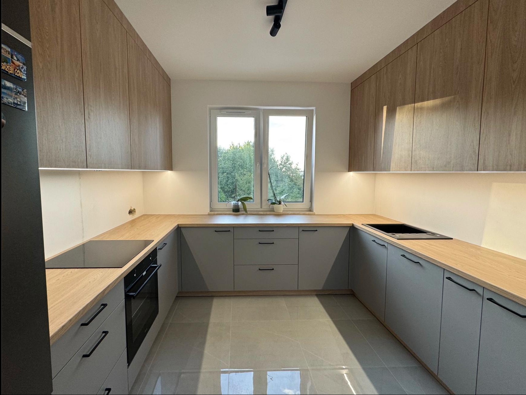 Nowoczesna kuchnia w kształcie litery U z szarymi szafkami i drewnianym blatem. Nad blatem okno z widokiem na zieleń. Oświetlenie LED podszafkowe.