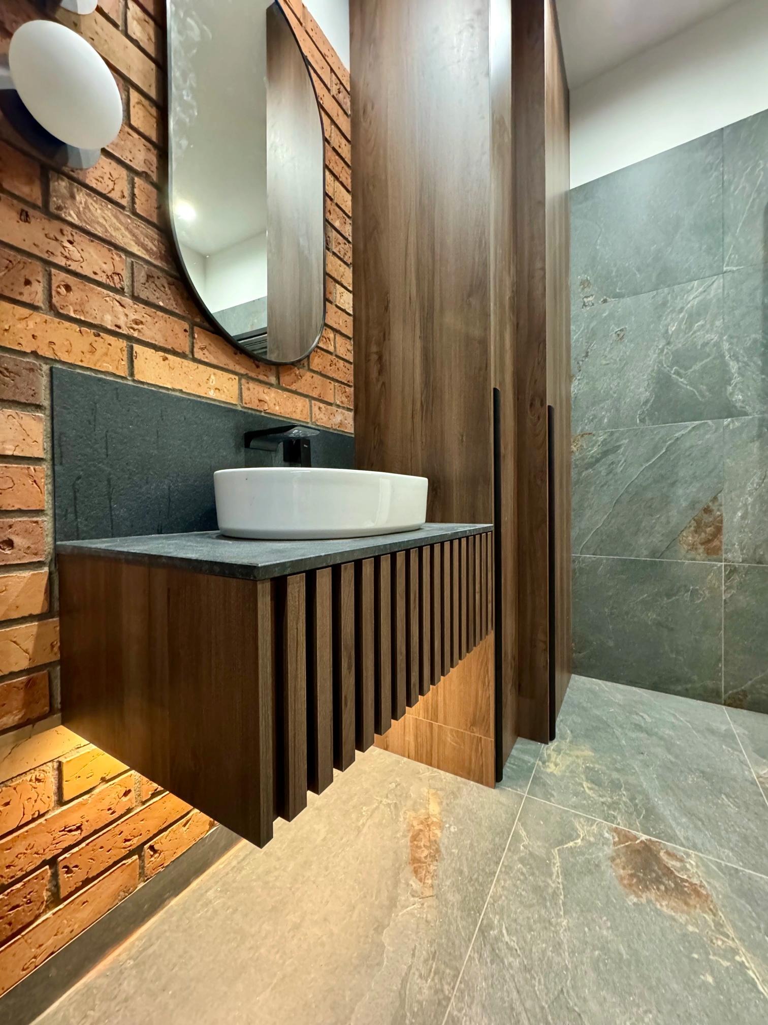 Nowoczesna łazienka z umywalką nablatową na szafce z lamelami, ceglaną ścianą i dużym lustrem. Widoczna zabudowa z drewnopodobnych płyt i płytki imitujące kamień.