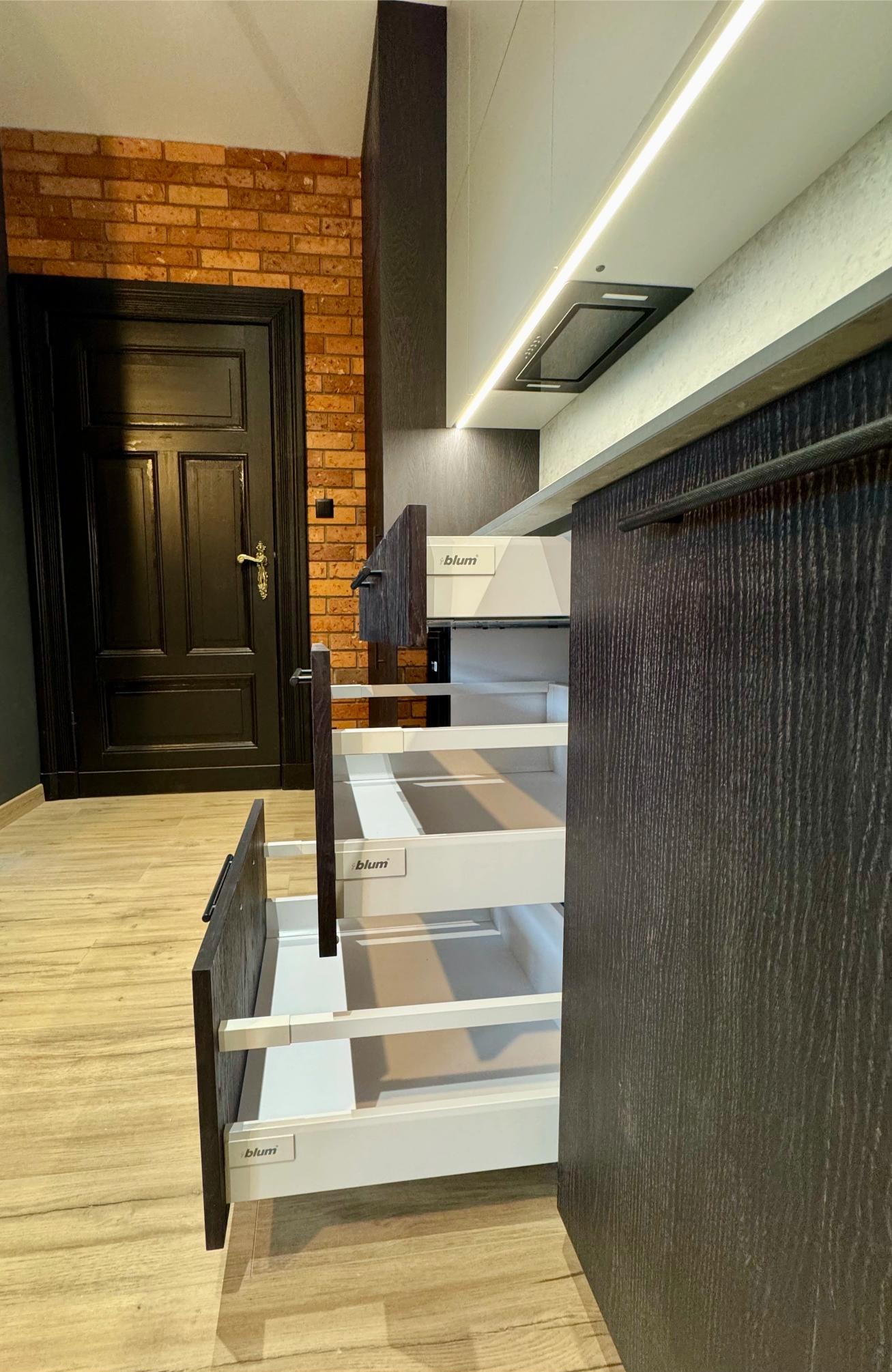 Nowoczesna kuchnia z szufladami Blum w kolorze ciemnego drewna, otwarte szuflady ukazują białe wnętrze. W tle ceglana ściana i czarne drzwi.