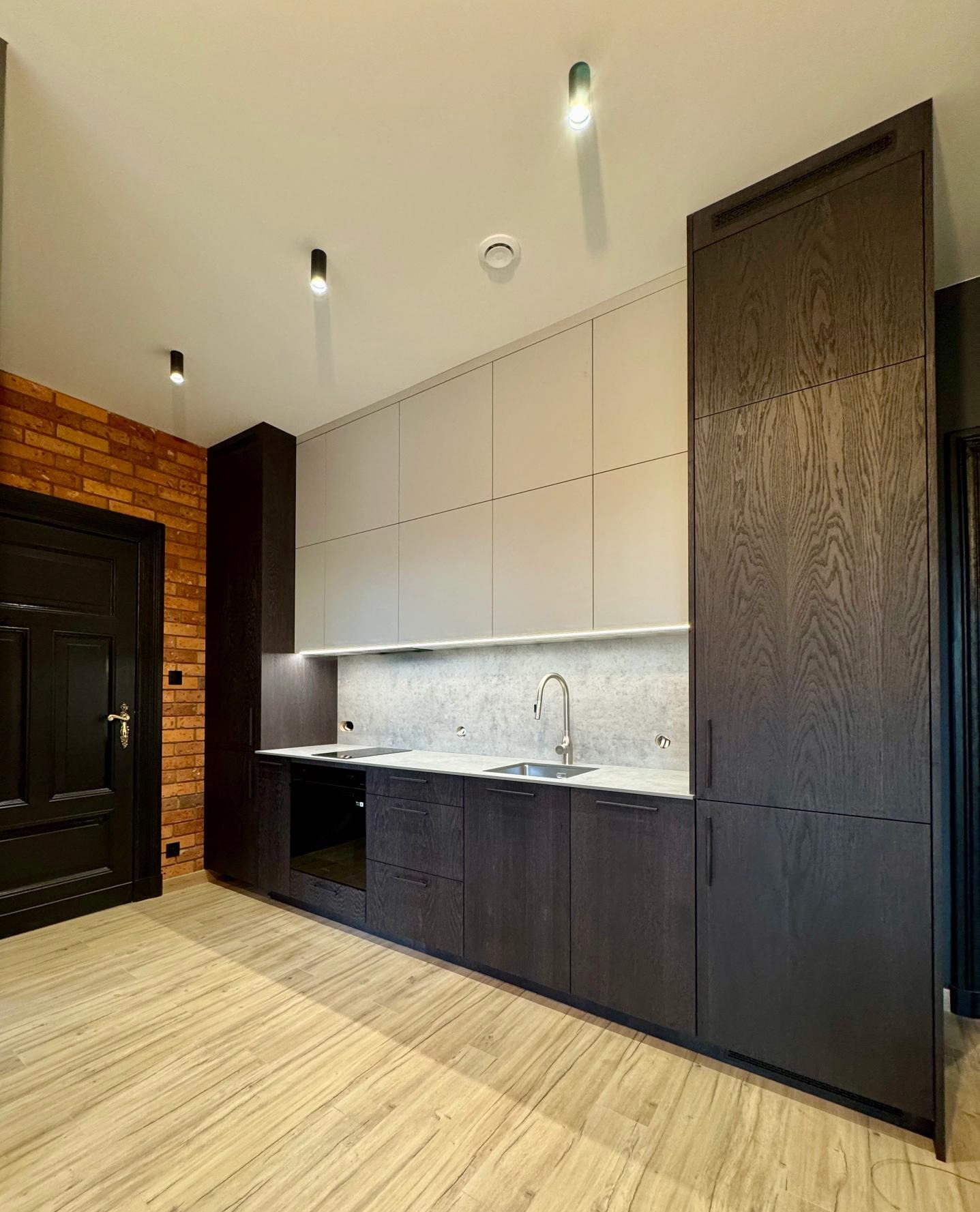 Nowoczesna kuchnia z ciemnymi drewnianymi szafkami, jasnymi górnymi szafkami i ceglaną ścianą z czarnymi drzwiami w tle. Oświetlenie LED pod szafkami.