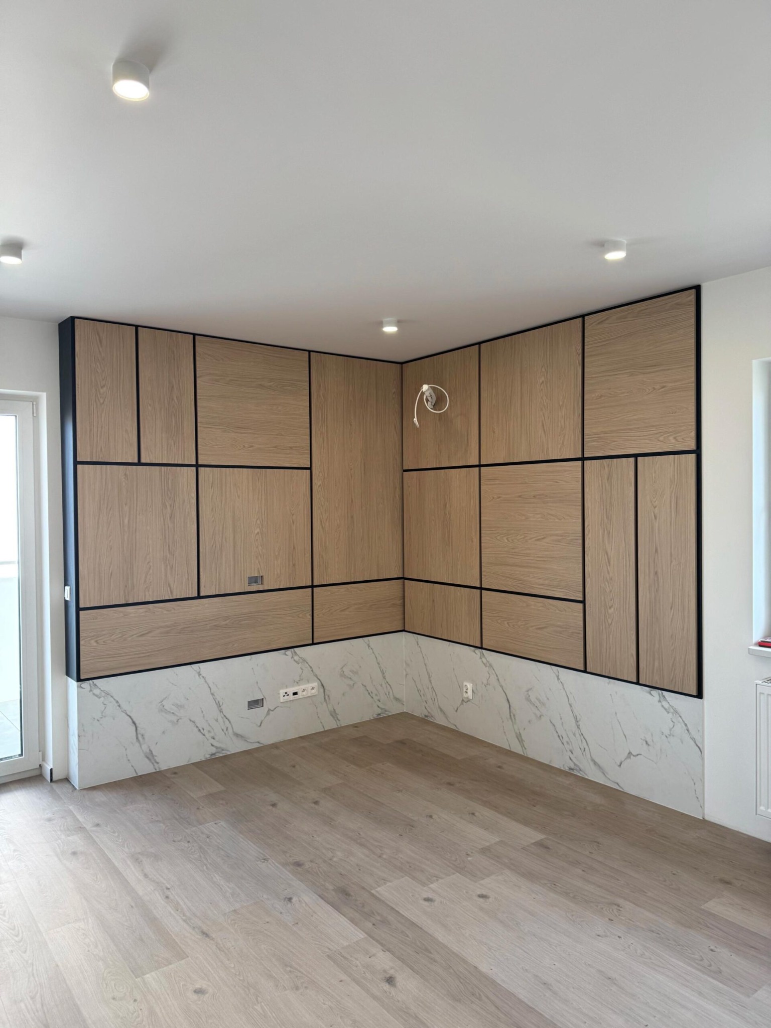 Nowoczesna aranżacja wnętrza z drewnianą ścianą panelową w geometryczne wzory, wykończoną marmurowym cokołem i podłogą w odcieniach drewna. Widoczne oświetlenie sufitowe.