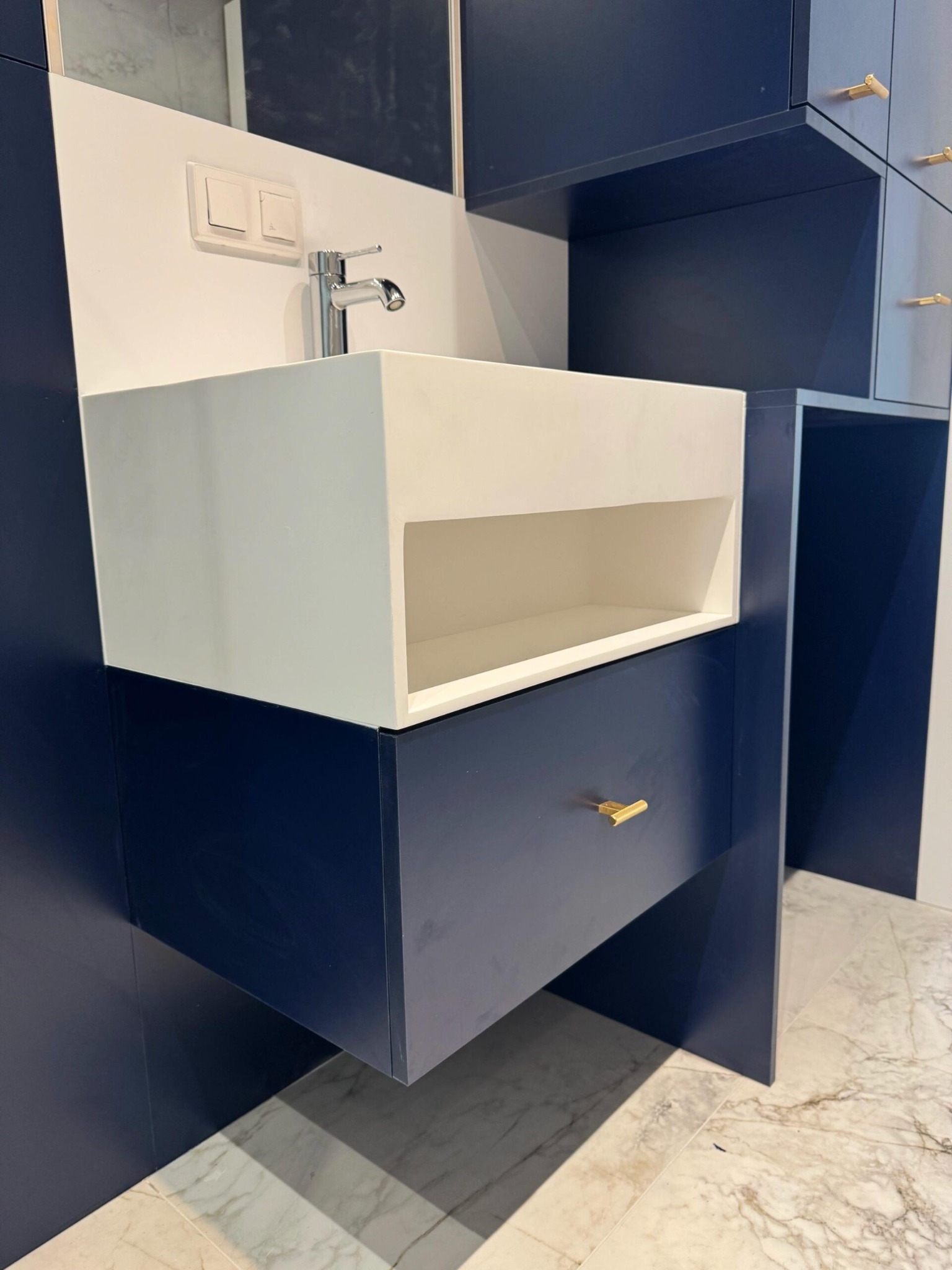 Nowoczesna, granatowo-biała umywalka z szafką i złotymi uchwytami, na tle marmurowej podłogi. Minimalistyczny design z widocznymi detalami armatury i oświetlenia.