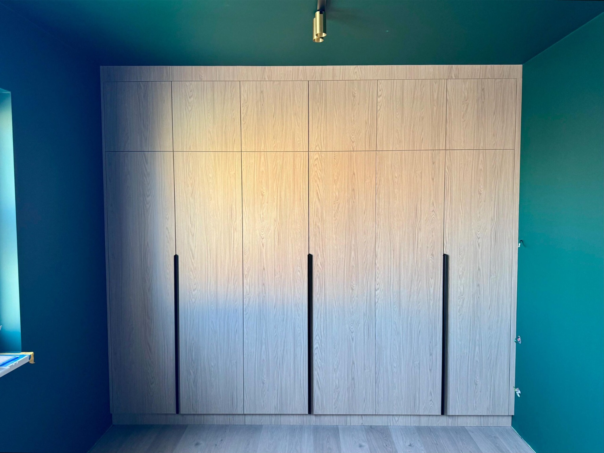 Jasna, drewnopodobna szafa wnękowa z czarnymi, pionowymi uchwytami, na tle ściany w kolorze morskim i szarej podłogi. Widoczne oświetlenie sufitowe.