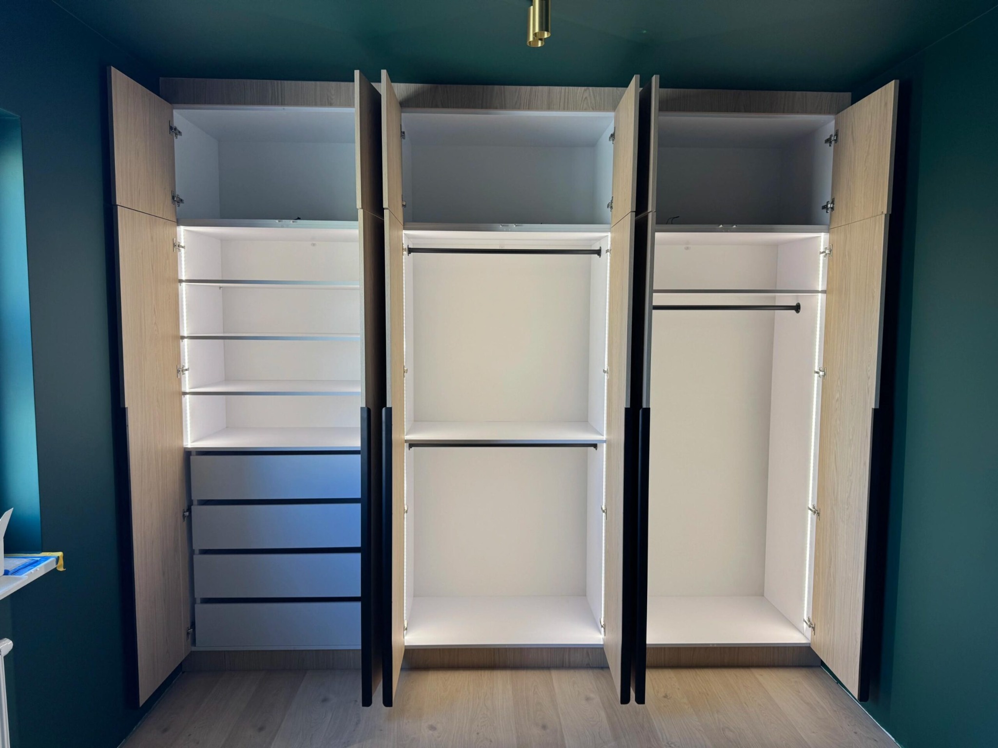 Otwarta szafa wnękowa z oświetleniem LED, widoczne półki, szuflady i drążek na ubrania. Jasne drewno frontów kontrastuje z ciemnozieloną ścianą.