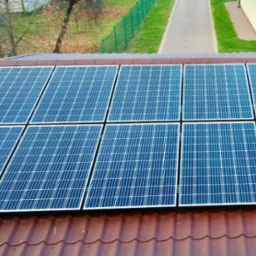 Panel słoneczny na brązowym dachu domu, widok z góry. Na tle zielona trawa i drzewa. Instalacja fotowoltaiczna na tle jesiennego krajobrazu.