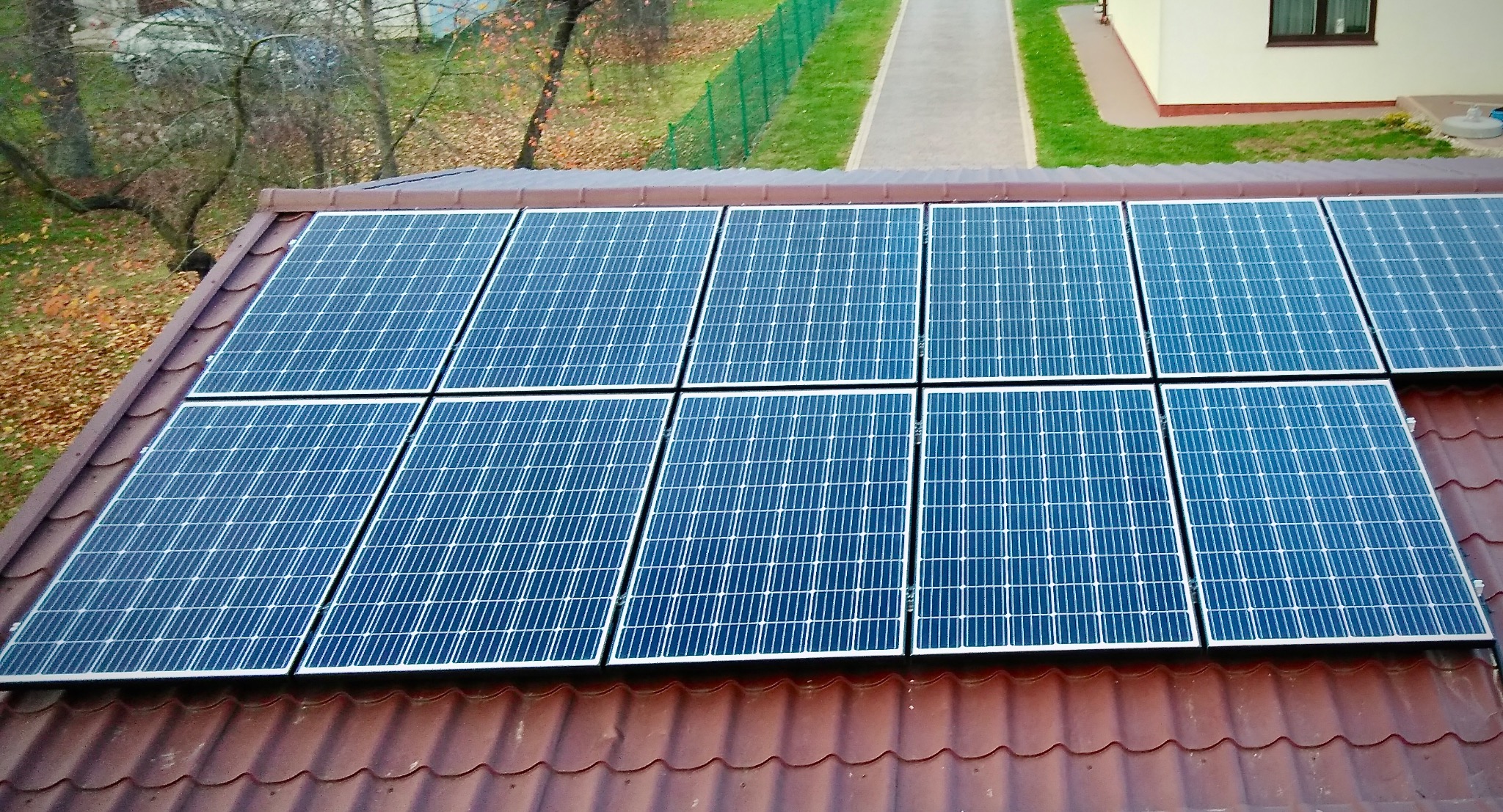 Panel słoneczny na brązowym dachu domu, widok z góry. Na tle zielona trawa i drzewa. Instalacja fotowoltaiczna na tle jesiennego krajobrazu.