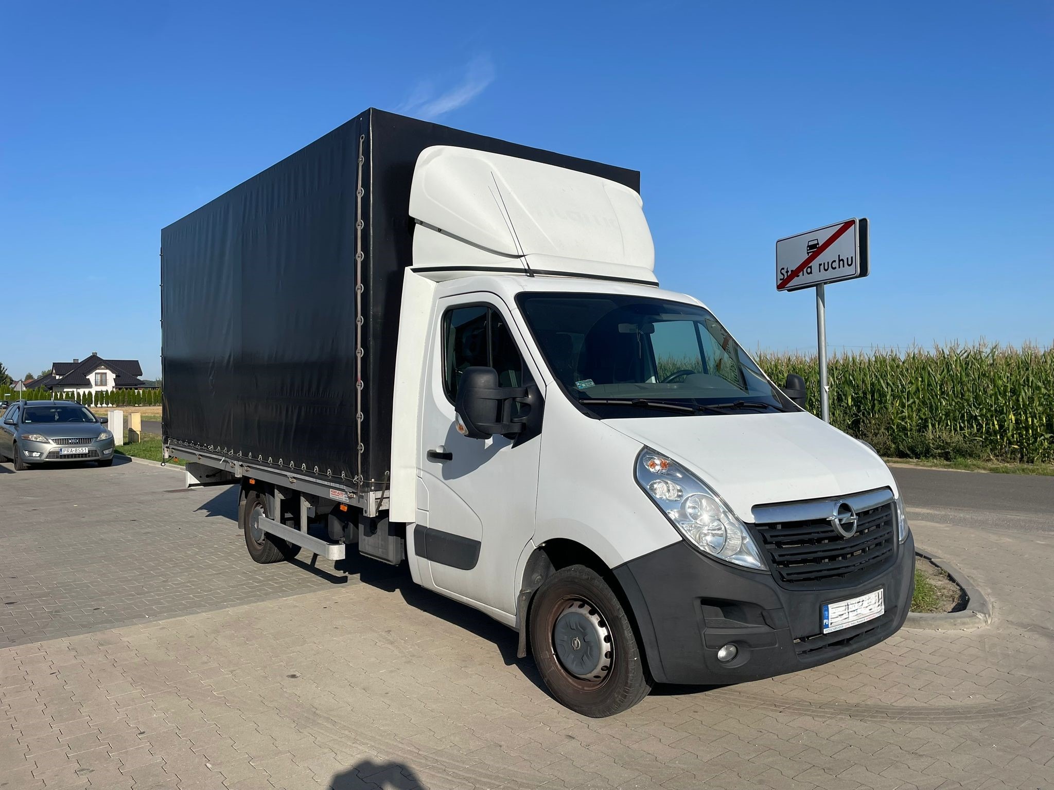 Biały bus Opel z czarną plandeką stoi na kostce brukowej obok znaku drogowego 'Strefa Ruchu'. W tle pole kukurydzy i dom.