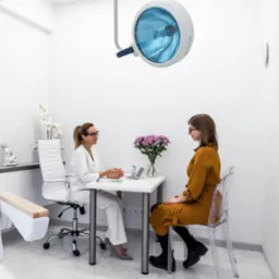 Kobieta w gabinecie lekarskim podczas konsultacji. Widoczny fotel zabiegowy, lampa operacyjna i kwiaty na stole.