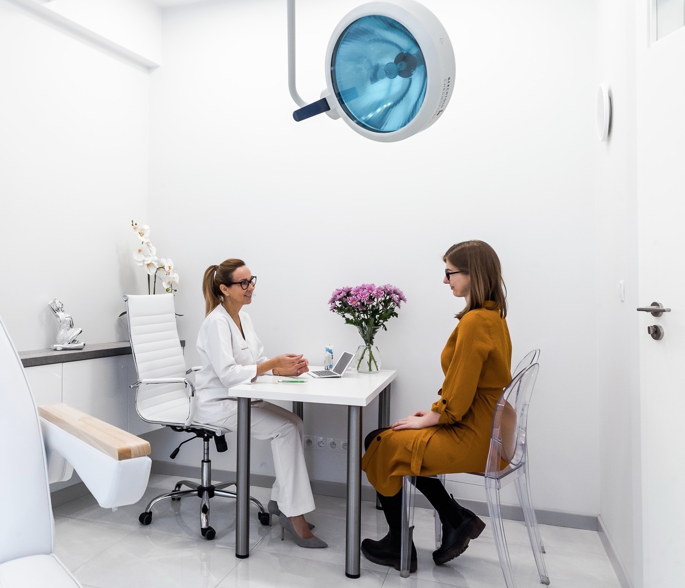 Kobieta w gabinecie lekarskim podczas konsultacji. Widoczny fotel zabiegowy, lampa operacyjna i kwiaty na stole.