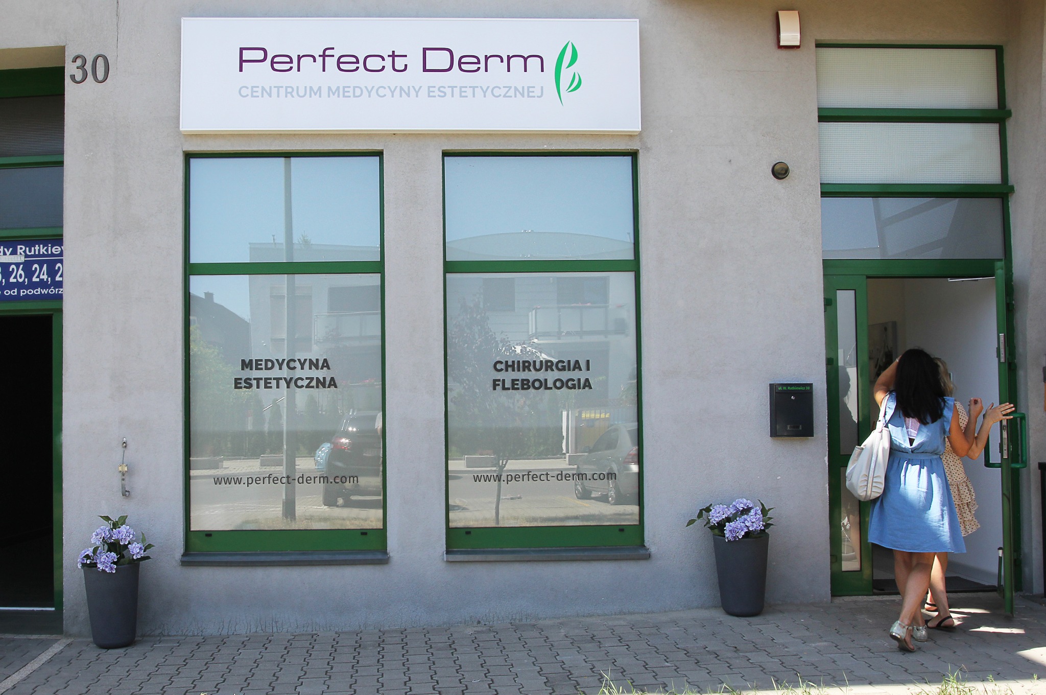 Wejście do centrum medycyny estetycznej Perfect Derm, z widocznymi oknami z napisem 'Medycyna Estetyczna' i 'Chirurgia i Flebologia'. Dwie kobiety wchodzą do środka.