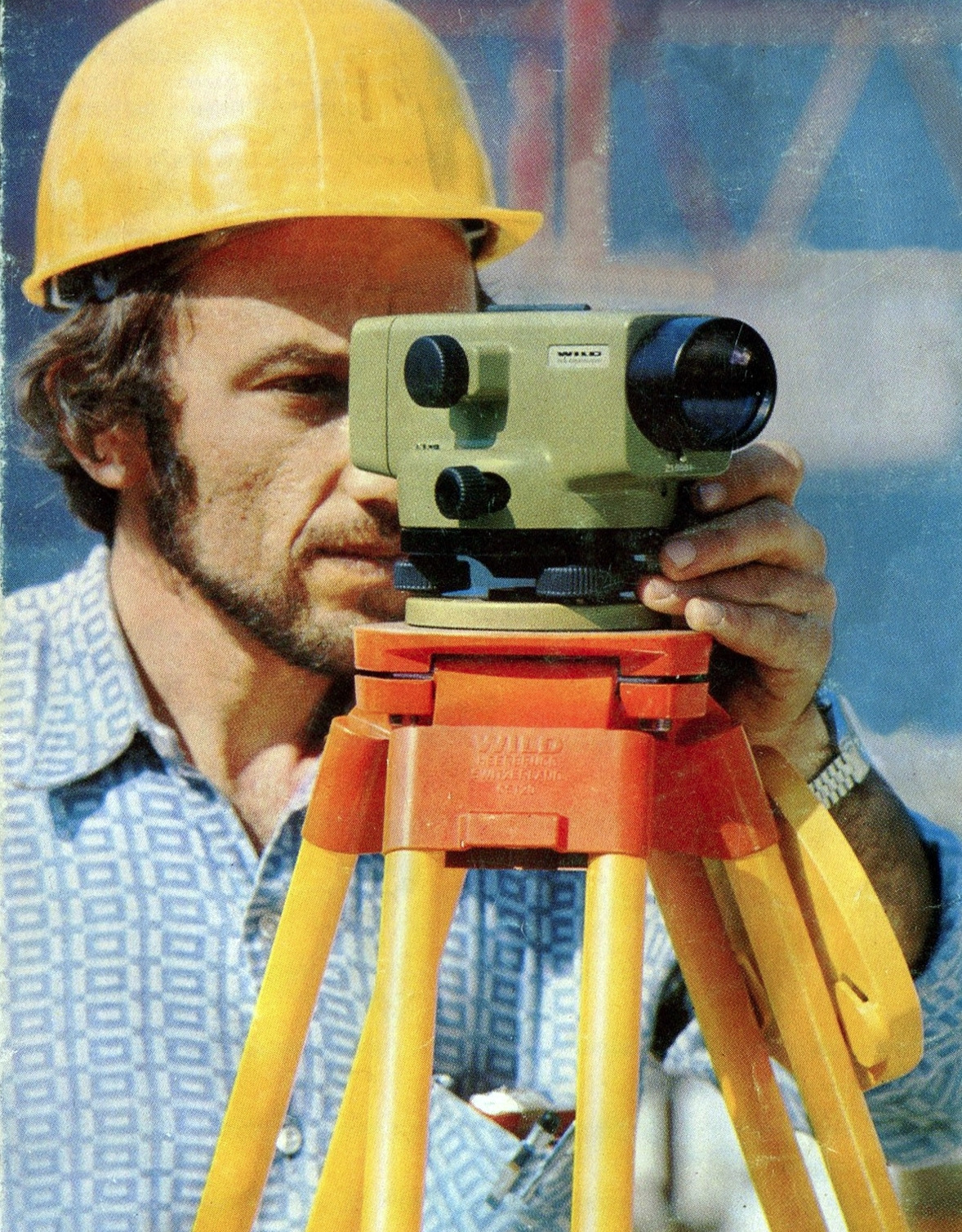 Mężczyzna w kasku, z brodą, celuje przez poziomicę Wild Heerbrugg na statywie, w tle błękitne niebo. Ujęcie z bliska na urządzenie i twarz.