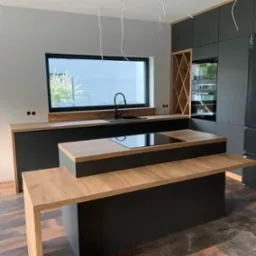 Nowoczesna kuchnia z wyspą i czarnymi szafkami, drewniany blat z płytą indukcyjną, okno z widokiem na ogród. Minimalistyczny design z elementami drewna.