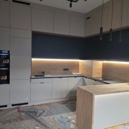 DSJ MEBLE - Nowoczesna kuchnia z wyspą: białe fronty szafek dolnych, czarne górne, drewniany panel nad blatem z oświetleniem LED. Na podłodze zabezpieczenia przed zabrudzeniem.