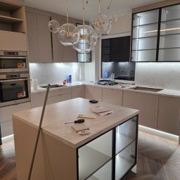 DSJ MEBLE - Montaż kuchni na wymiar: wyspa z marmurowym blatem, szklane witryny, oświetlenie LED pod szafkami, wiszące lampy kuliste. Widoczne narzędzia i dokumenty na blacie.