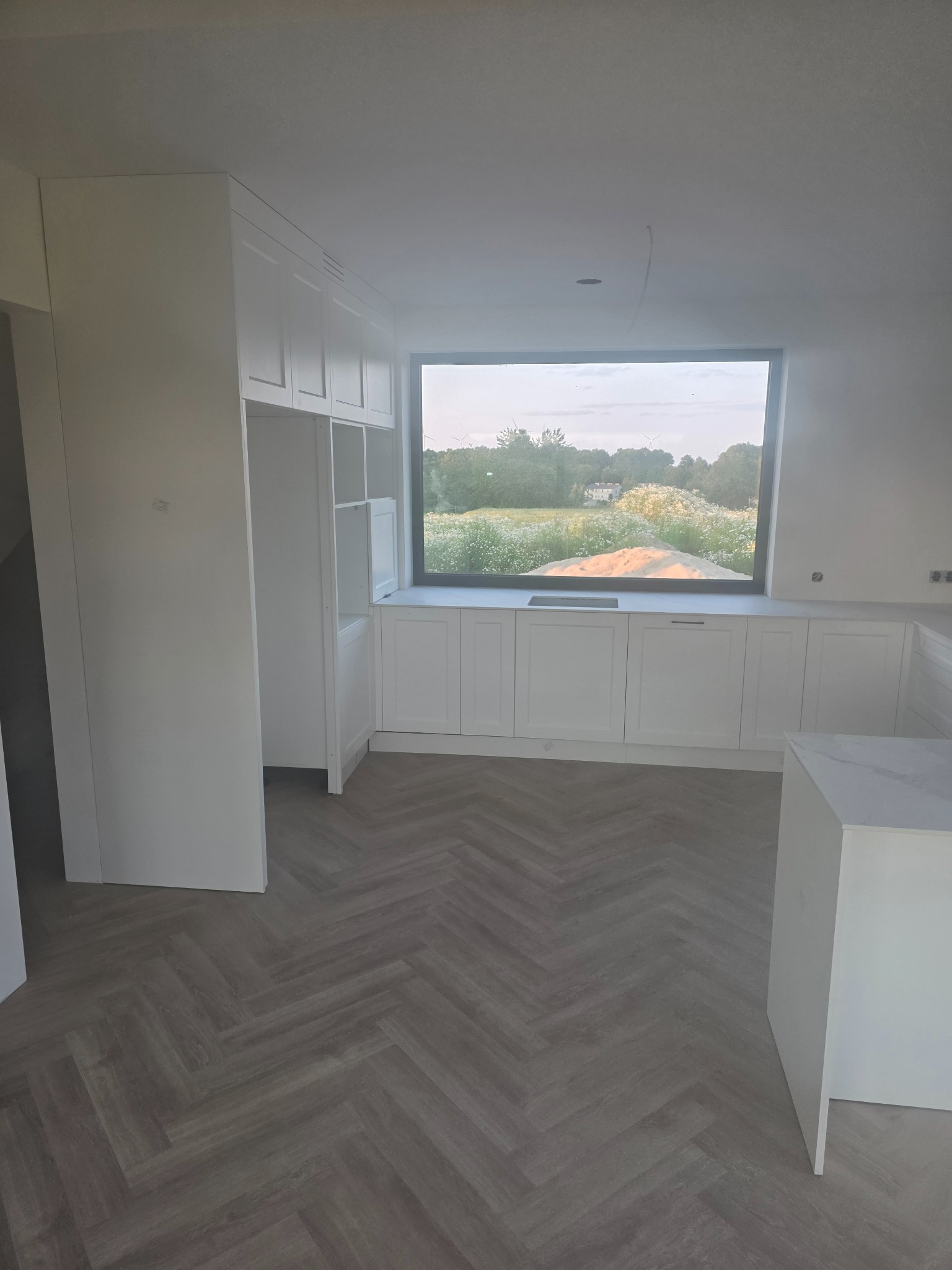 Jasna kuchnia na wymiar z oknem na ogród, szafki z białymi frontami, parkiet w jodełkę, minimalistyczny design.