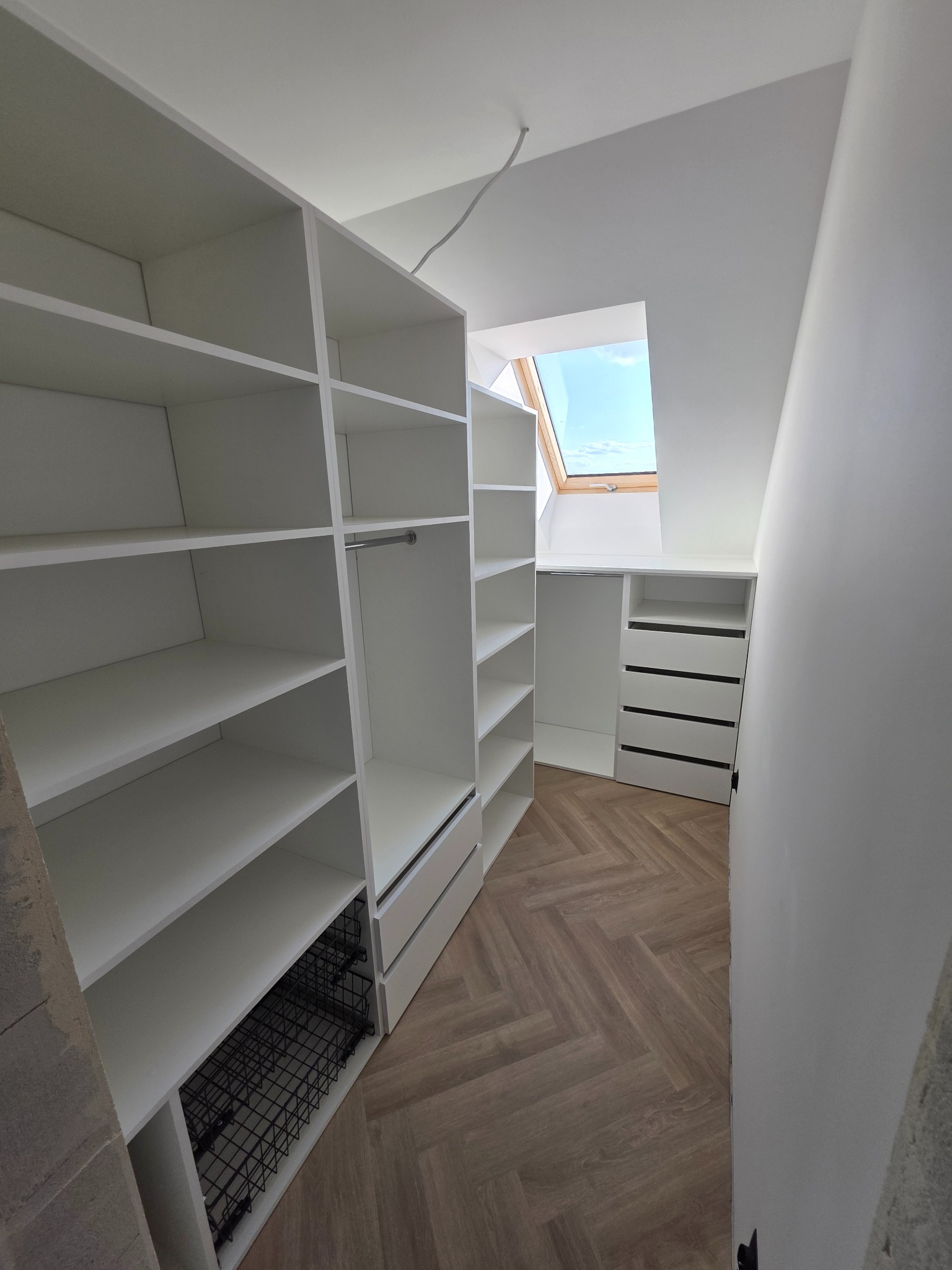 Biała, przestronna garderoba na poddaszu z oknem dachowym. Widoczne półki, szuflady, drążek na ubrania i kosz cargo. Podłoga w jodełkę.