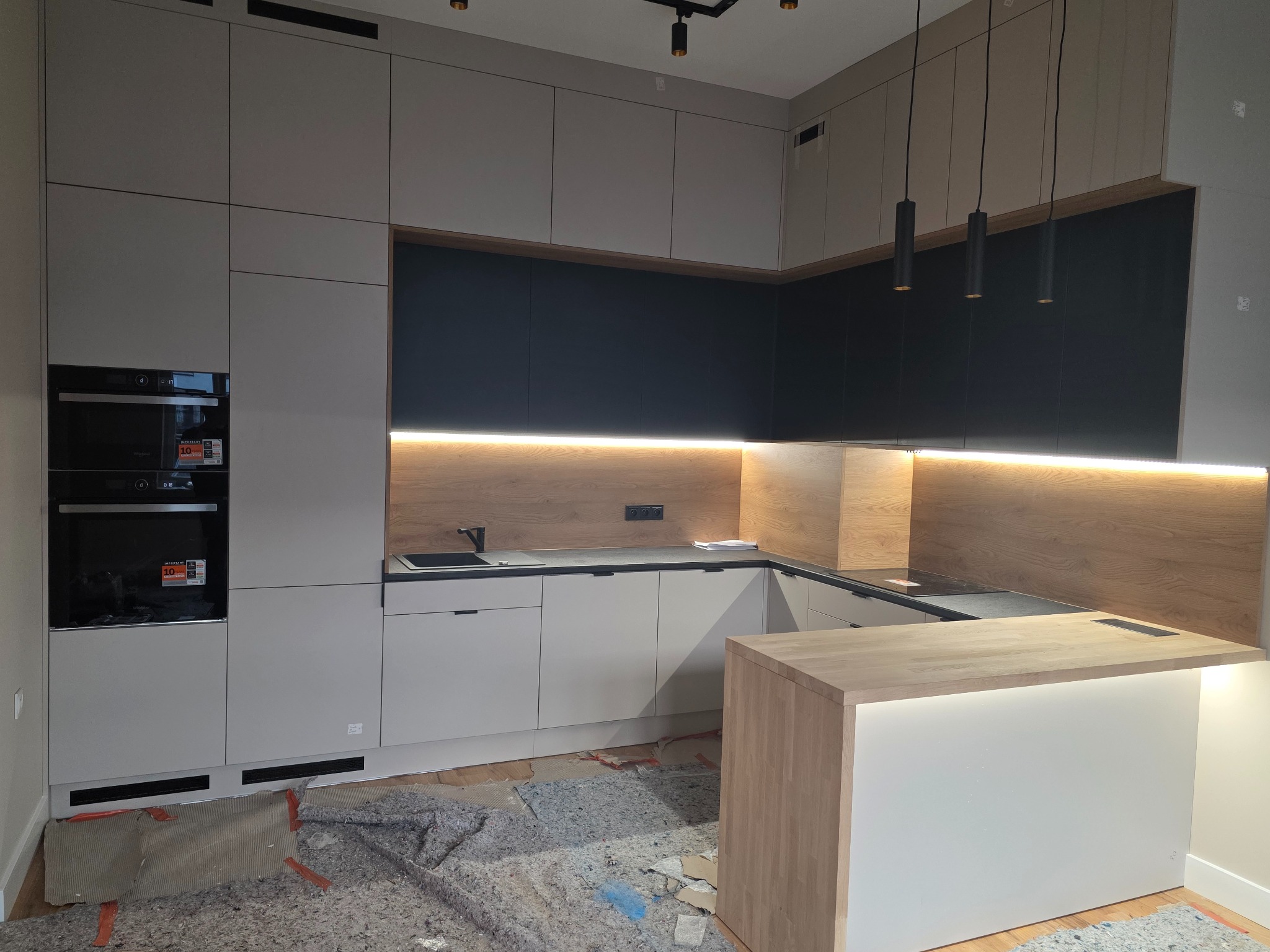 Nowoczesna kuchnia z wyspą: białe fronty szafek dolnych, czarne górne, drewniany panel nad blatem z oświetleniem LED. Na podłodze zabezpieczenia przed zabrudzeniem.