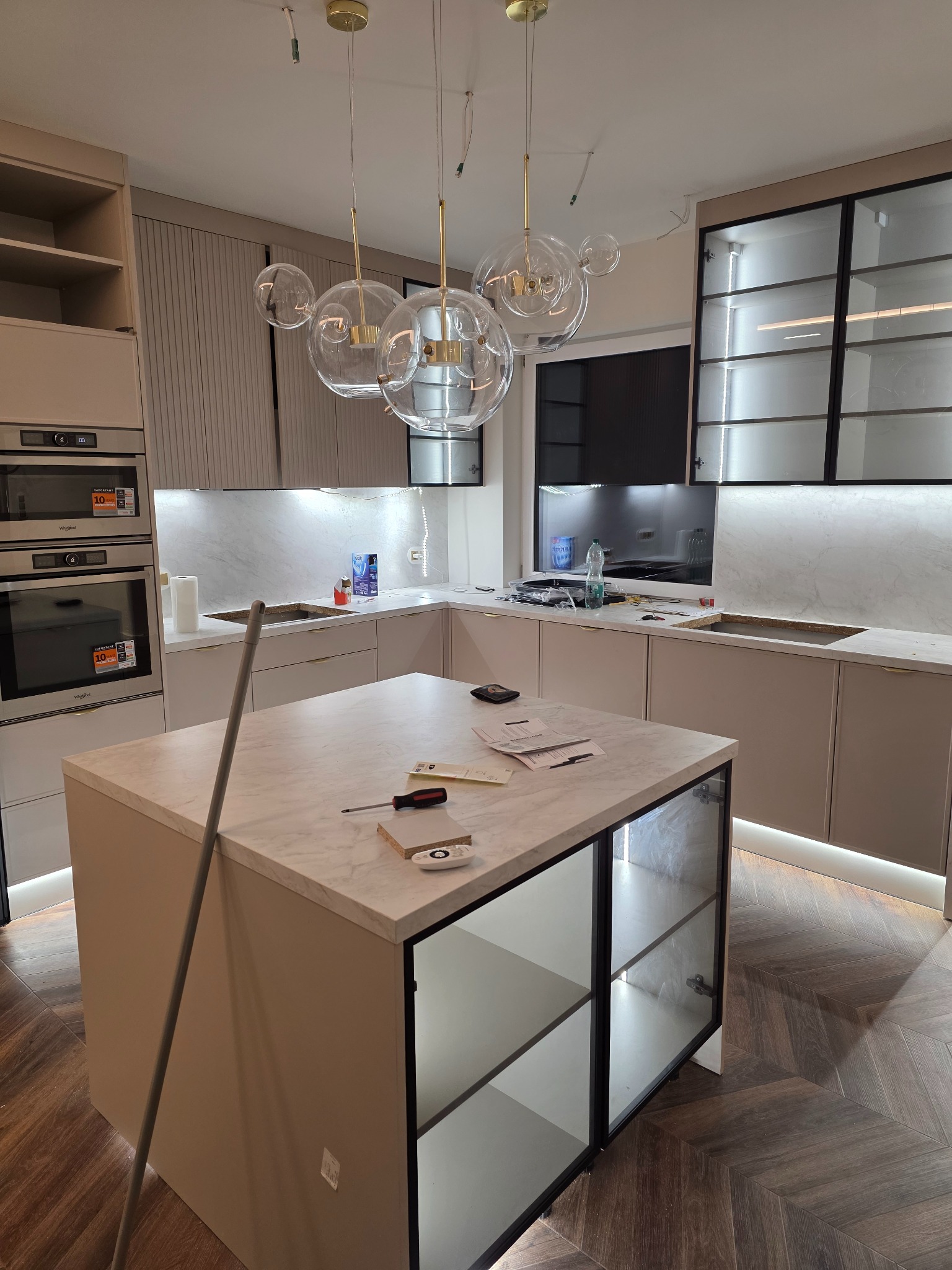 Montaż kuchni na wymiar: wyspa z marmurowym blatem, szklane witryny, oświetlenie LED pod szafkami, wiszące lampy kuliste. Widoczne narzędzia i dokumenty na blacie.
