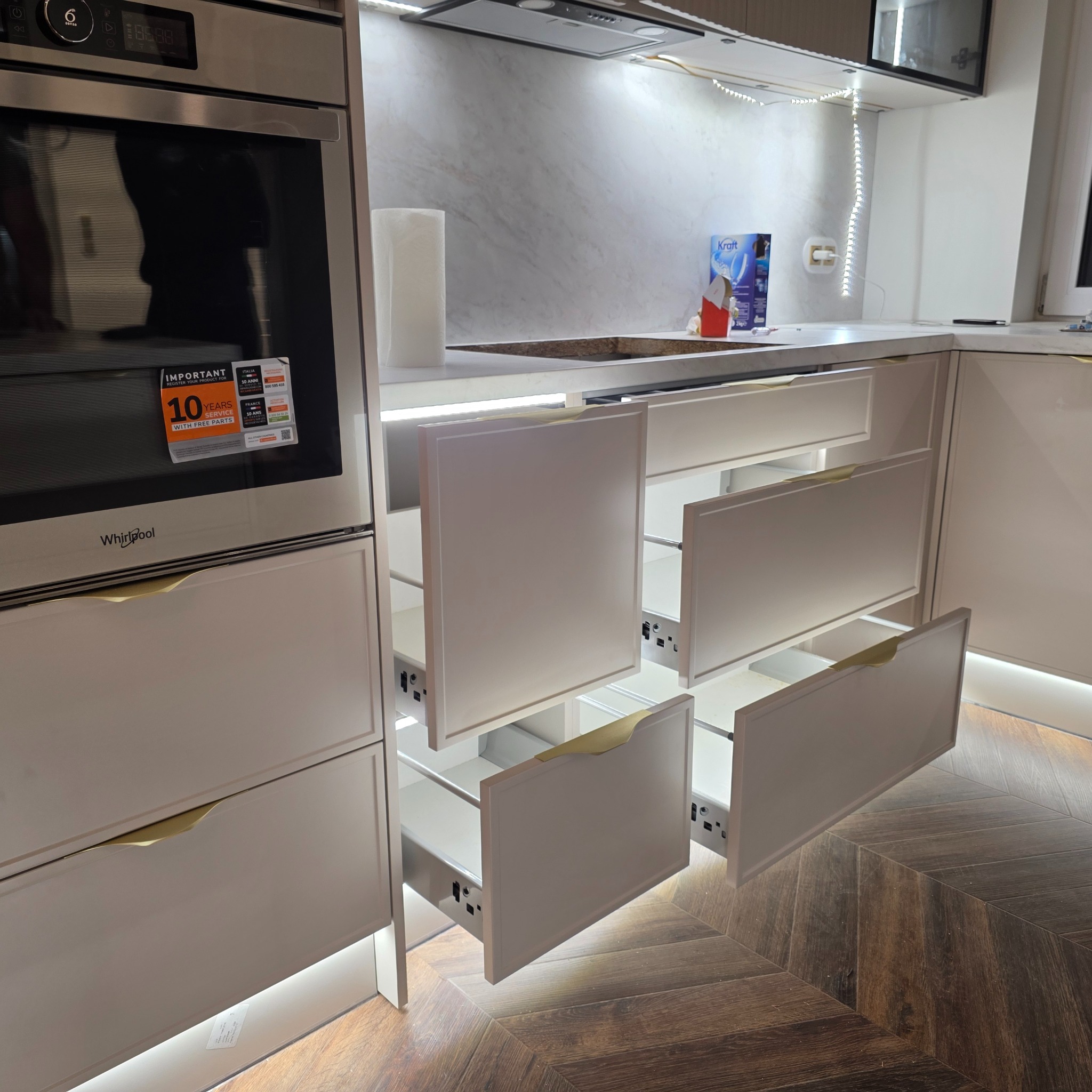 Nowoczesna kuchnia z białymi frontami i złotymi uchwytami, oświetlenie LED pod szafkami, otwarte szuflady z widocznym mechanizmem, parkiet w jodełkę.