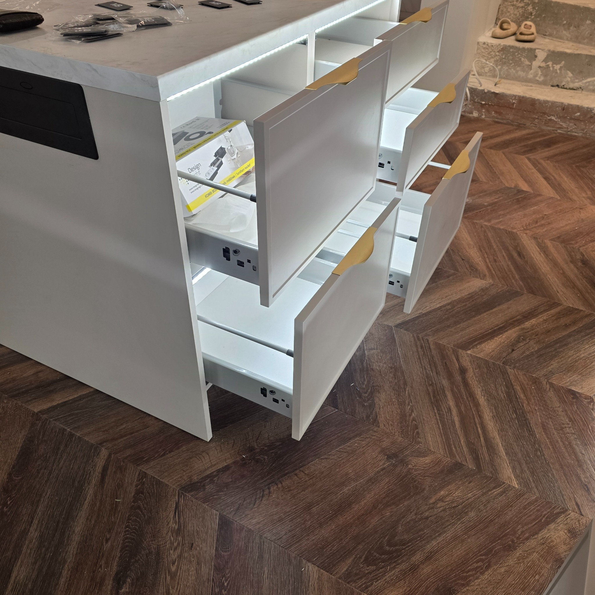 Biała wyspa kuchenna z otwartymi szufladami, oświetleniem LED i złotymi uchwytami, na tle parkietu w jodełkę i schodów. Widoczny osprzęt.