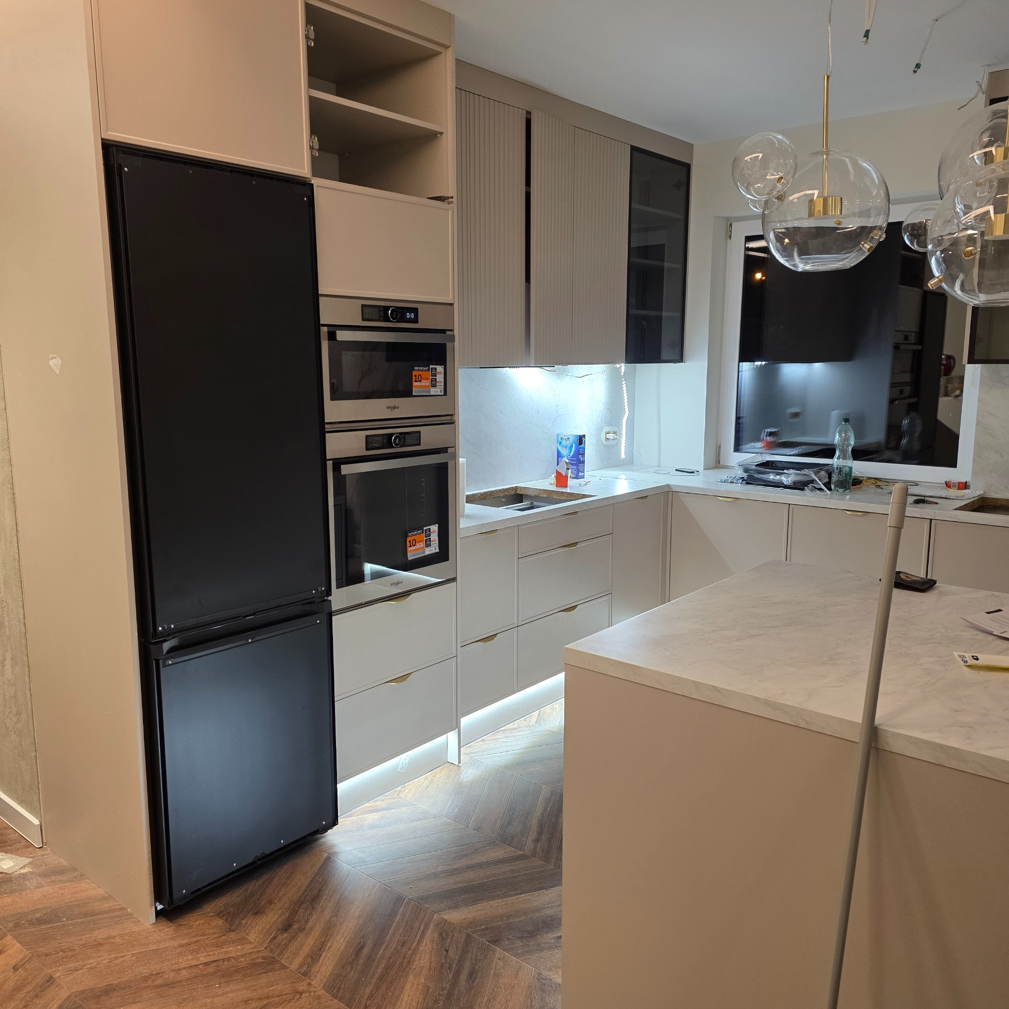 Jasna kuchnia z wyspą, fronty szafek z subtelnymi żłobieniami, oświetlenie LED pod szafkami, czarna lodówka w zabudowie, parkiet w jodełkę.