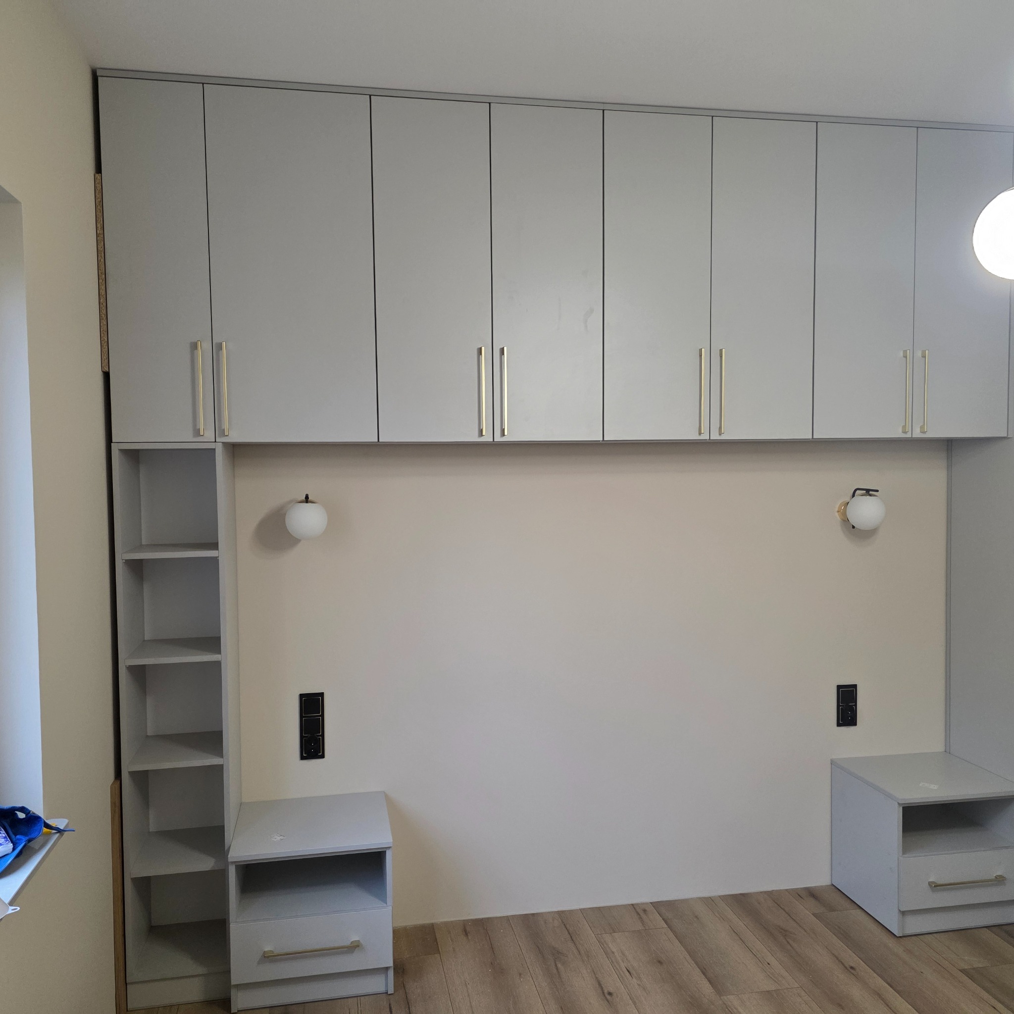 Jasnoszara szafa wnękowa z szafkami górnymi i otwartym regałem, dopasowana do szafek nocnych. Złote uchwyty, drewnopodobna podłoga.