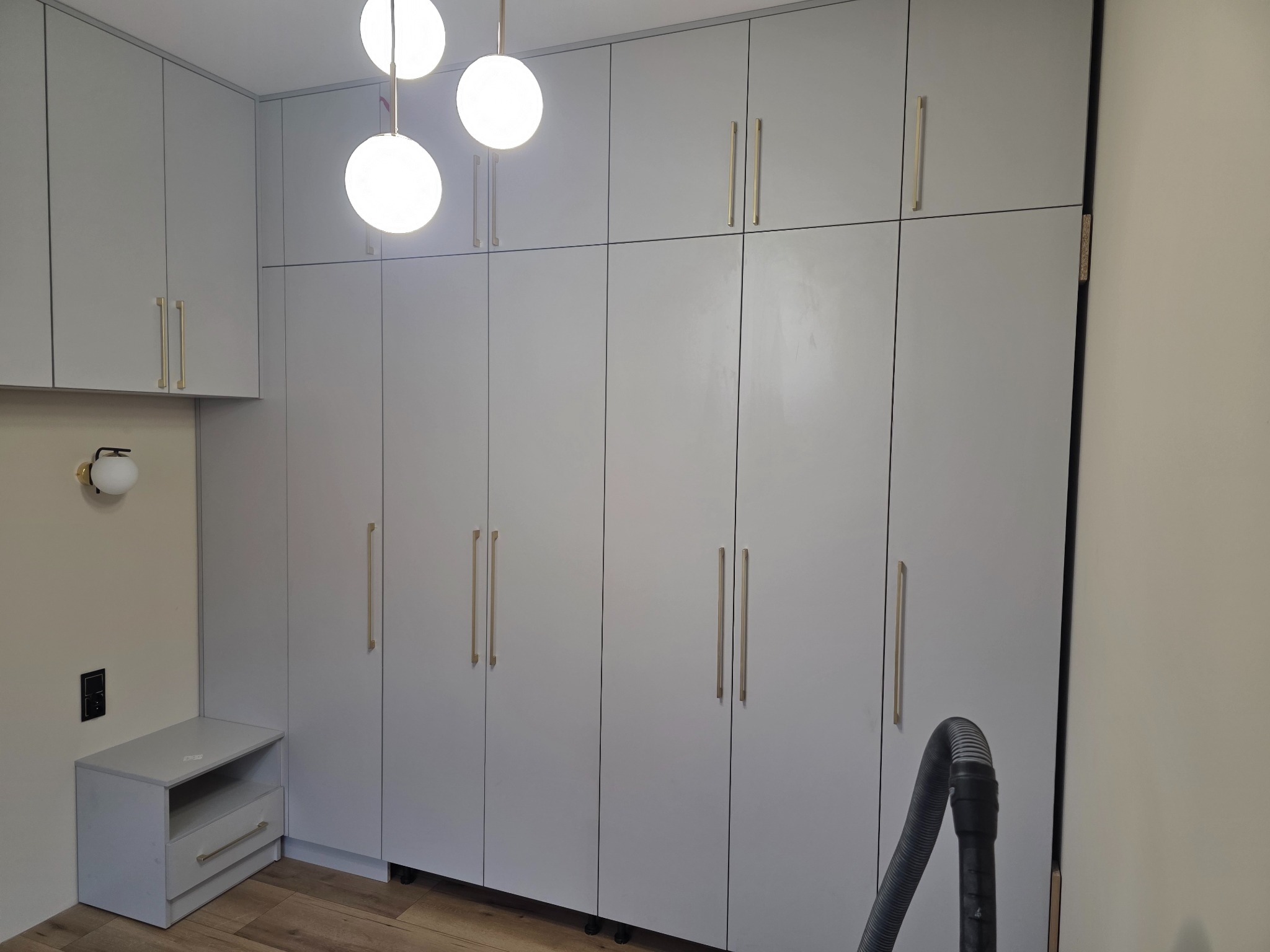 Jasnoszara zabudowa na wymiar z podświetleniem i złotymi uchwytami, uzupełniona szafką nocną. Widoczny fragment odkurzacza.