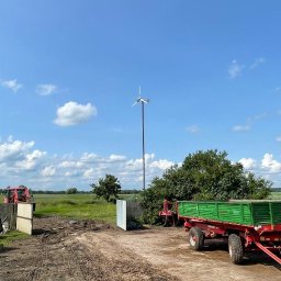 Green Moon Energy Sp. z o.o. - Samodzielna turbina wiatrowa na polu uprawnym, z traktorem i przyczepą w tle, pod błękitnym niebem z chmurami. Energetyka wiatrowa na wsi.
