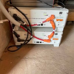 Green Moon Energy Sp. z o.o. - Dwa białe moduły bateryjne litowo-jonowe marki Deye, jeden na drugim, z kablami i przełącznikami, w pomieszczeniu ze ścianą z cegły i betonową podłogą.