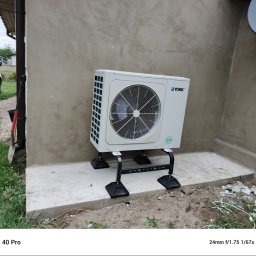 Green Moon Energy Sp. z o.o. - Biała pompa ciepła York, model R32, zamontowana na zewnątrz budynku o szarej elewacji, widoczna antena satelitarna. Częściowo widoczny pracownik i samochód.