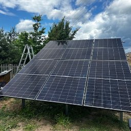 Green Moon Energy Sp. z o.o. - Panel słoneczny na trawie, drabina oparta o konstrukcję. Tło: ogród, drewniany płot, niebo z chmurami. Widoczne narzędzia i skrzynka narzędziowa.