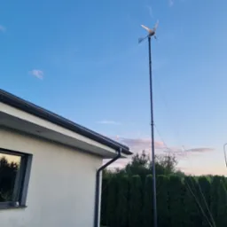 Mała turbina wiatrowa przy domu jednorodzinnym, widoczna na tle błękitnego nieba o zmierzchu. Ujęcie z dołu, z perspektywy ogrodu.