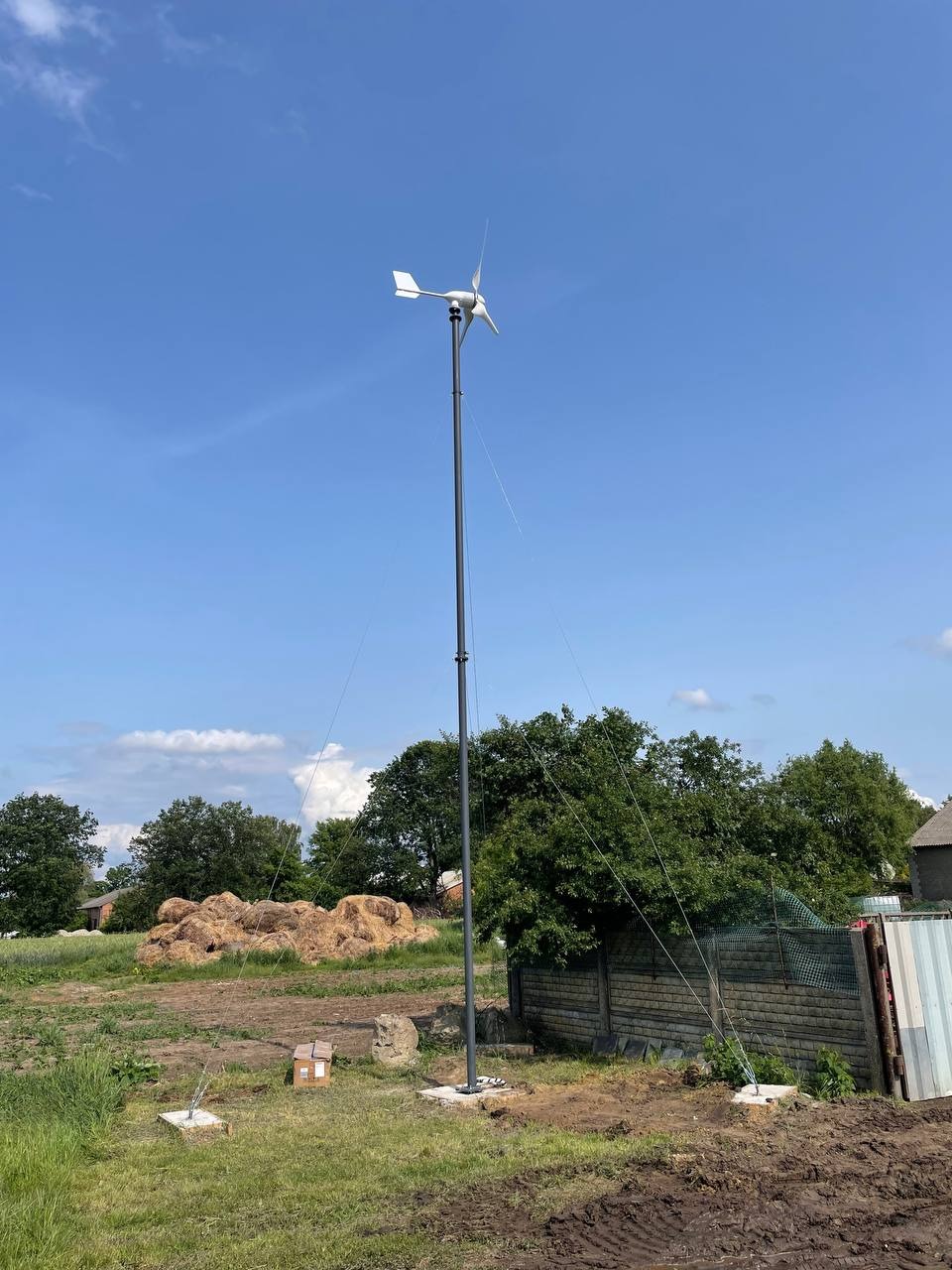 Wysoka turbina wiatrowa na tle błękitnego nieba, zamontowana na działce z widocznym stogiem siana i ogrodzeniem. Ujęcie z dołu podkreśla wysokość instalacji.