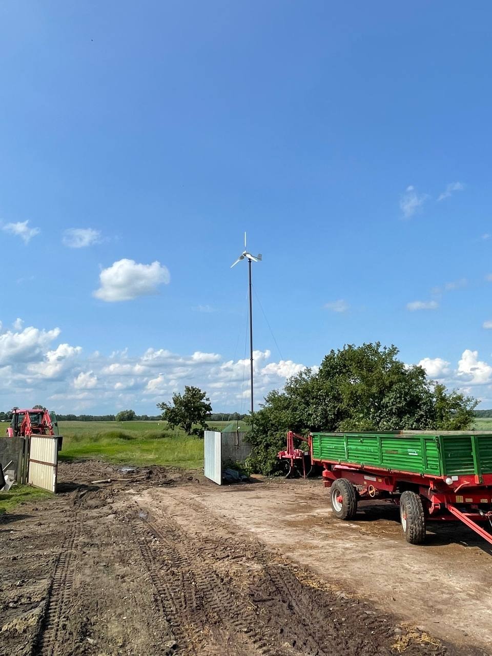 Samodzielna turbina wiatrowa na polu uprawnym, z traktorem i przyczepą w tle, pod błękitnym niebem z chmurami. Energetyka wiatrowa na wsi.