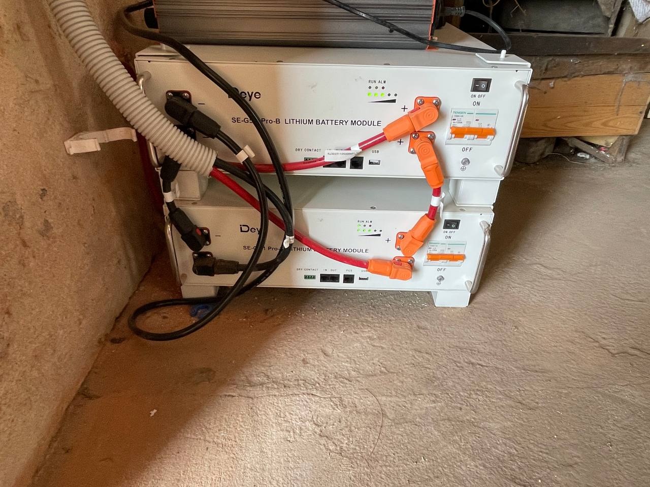 Dwa białe moduły bateryjne litowo-jonowe marki Deye, jeden na drugim, z kablami i przełącznikami, w pomieszczeniu ze ścianą z cegły i betonową podłogą.