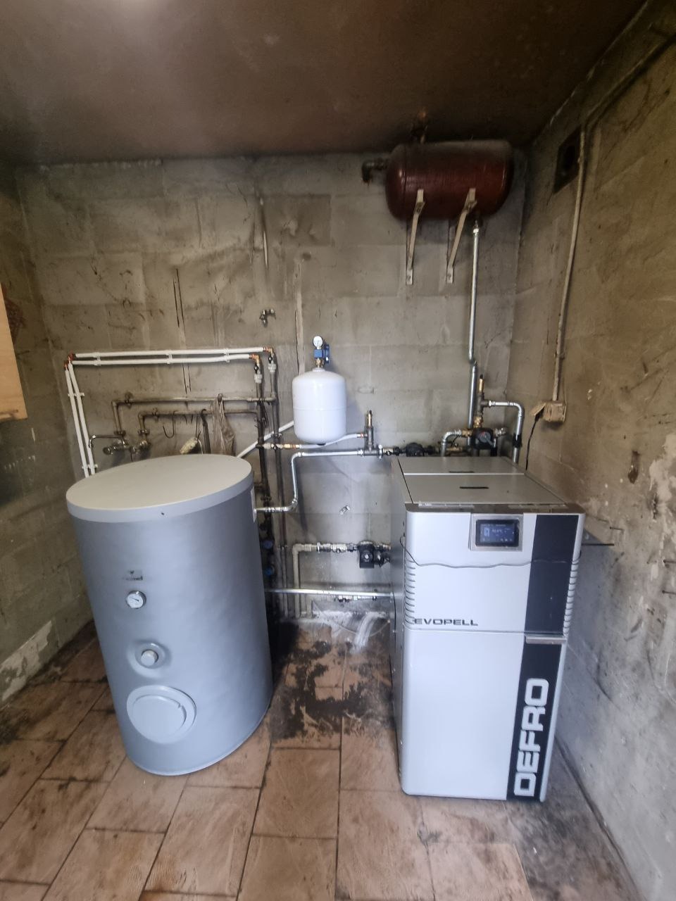 Instalacja kotła DEFRO Evopell z zasobnikiem ciepłej wody w pomieszczeniu z surowymi ścianami i brązowym zbiornikiem wyrównawczym na górze.