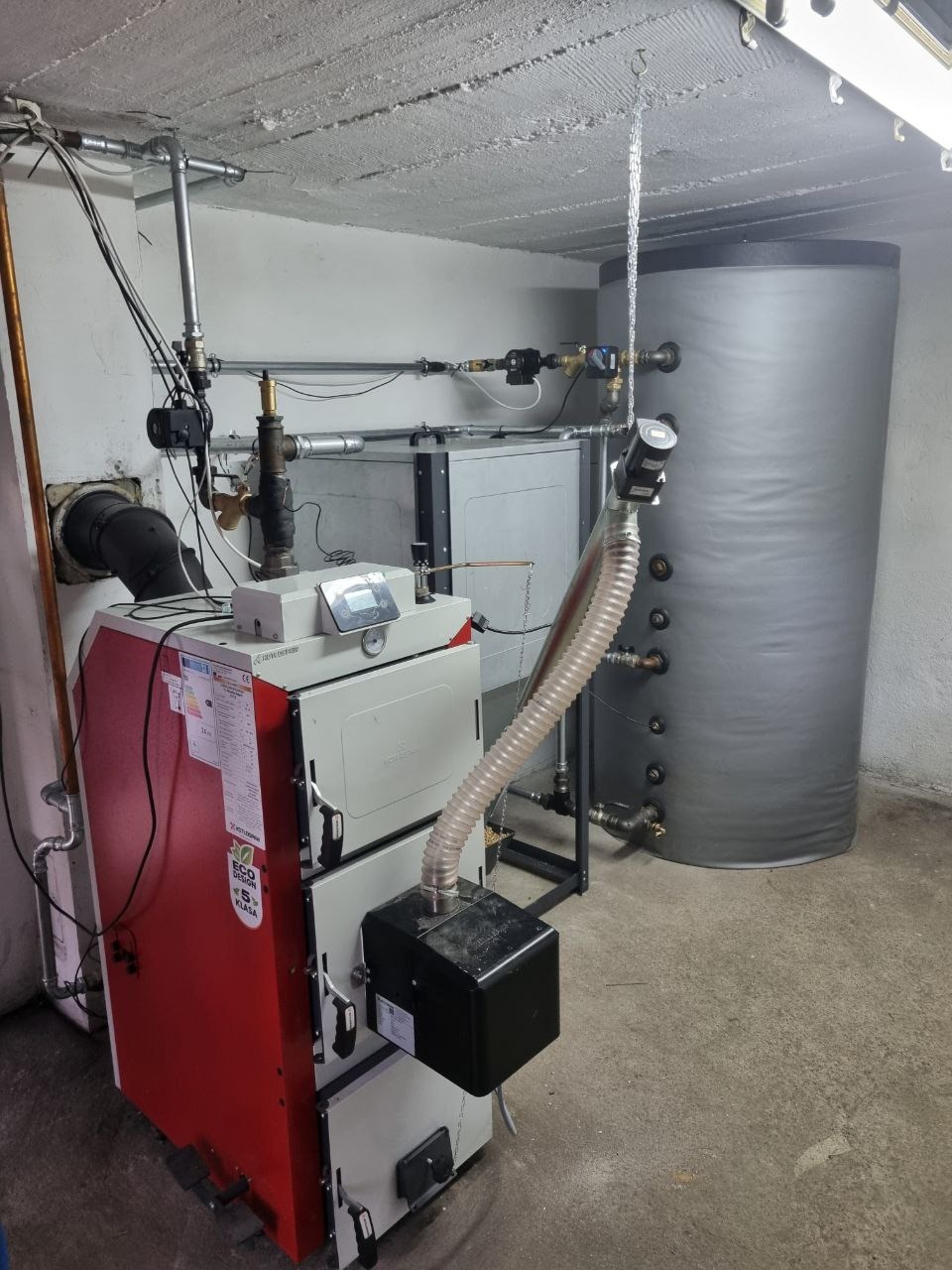 Kocioł na pellet z podajnikiem i zasobnikiem wody w kotłowni. Instalacja grzewcza z osprzętem. Widoczne rury, zawory, sterownik i zbiornik buforowy.