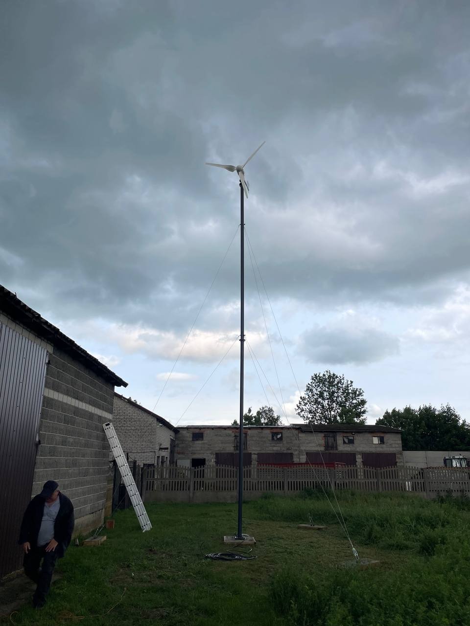 Mała turbina wiatrowa na podwórku, wsparta linami, z budynkami w tle i pochmurnym niebem. Osoba w oddali, drabina oparta o budynek.