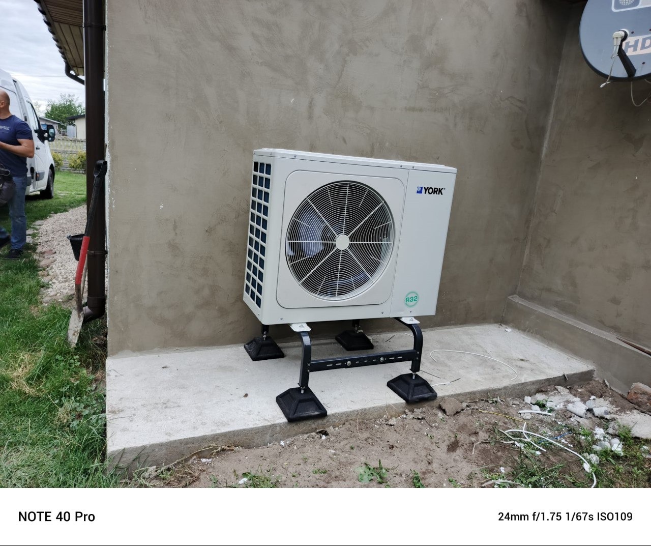 Biała pompa ciepła York, model R32, zamontowana na zewnątrz budynku o szarej elewacji, widoczna antena satelitarna. Częściowo widoczny pracownik i samochód.