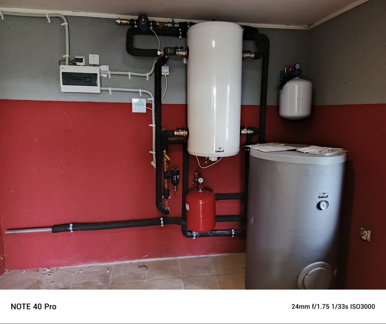 Instalacja grzewcza z dwoma zbiornikami i rurami na czerwono-szarej ścianie. Widoczne elementy automatyki i zabezpieczeń. Montaż w pomieszczeniu technicznym.