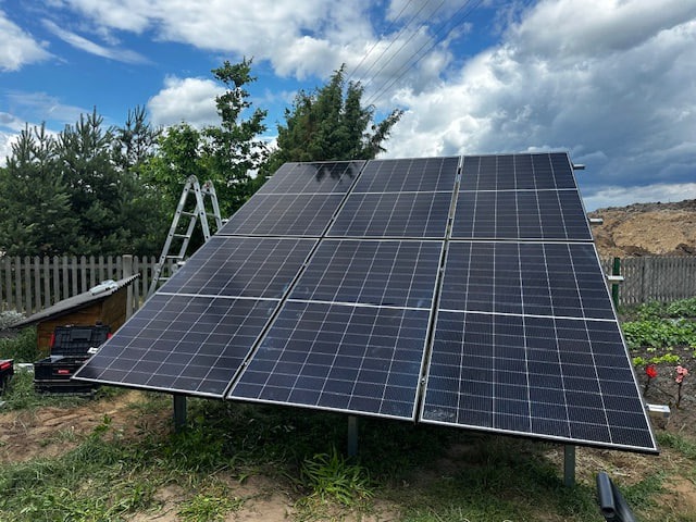 Panel słoneczny na trawie, drabina oparta o konstrukcję. Tło: ogród, drewniany płot, niebo z chmurami. Widoczne narzędzia i skrzynka narzędziowa.