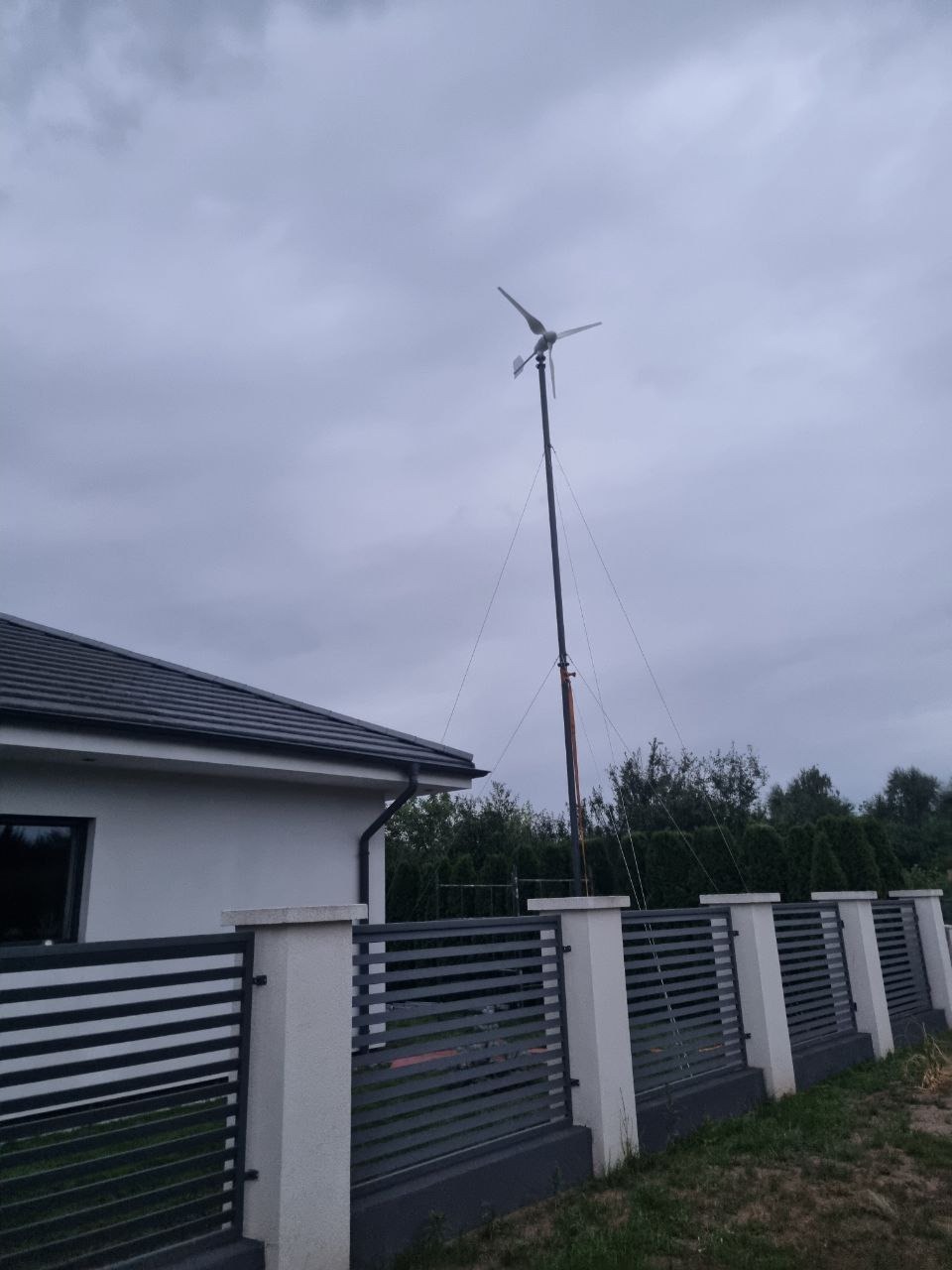 Turbina wiatrowa na posesji z szarym ogrodzeniem panelowym i białym domem pod pochmurnym niebem. Maszt turbiny podtrzymywany przez liny.
