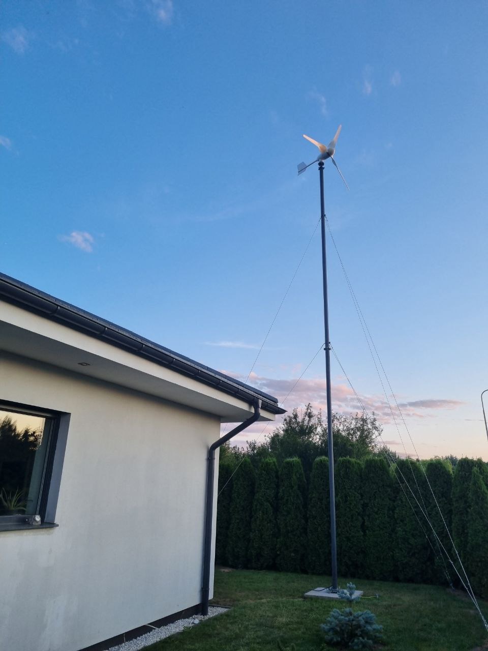 Mała turbina wiatrowa przy domu jednorodzinnym, widoczna na tle błękitnego nieba o zmierzchu. Ujęcie z dołu, z perspektywy ogrodu.