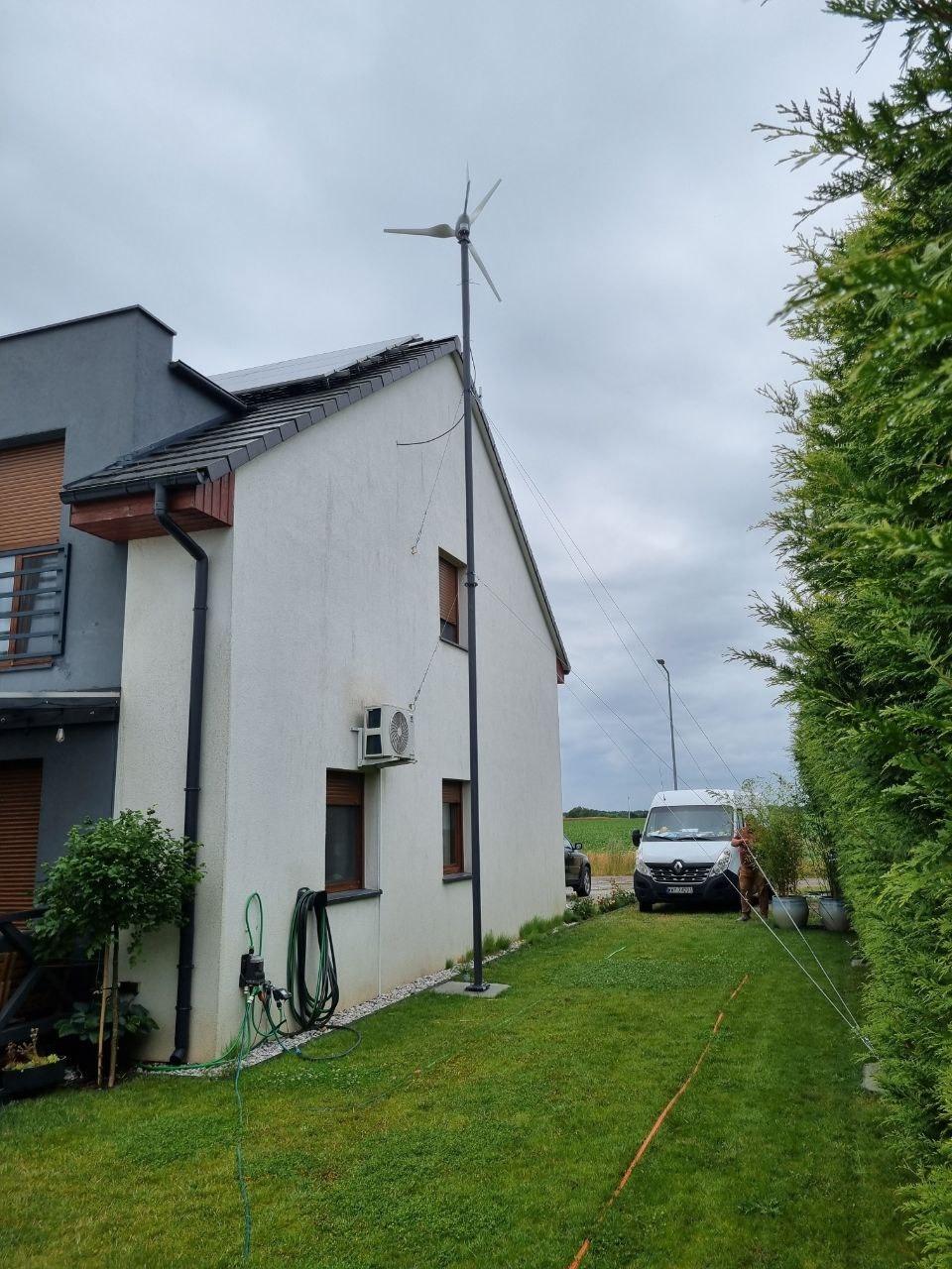 Dom z turbiną wiatrową na dachu i panelami solarnymi, widok z ogrodu, biała fasada, szara dobudówka. Na trawniku stoi biały bus.