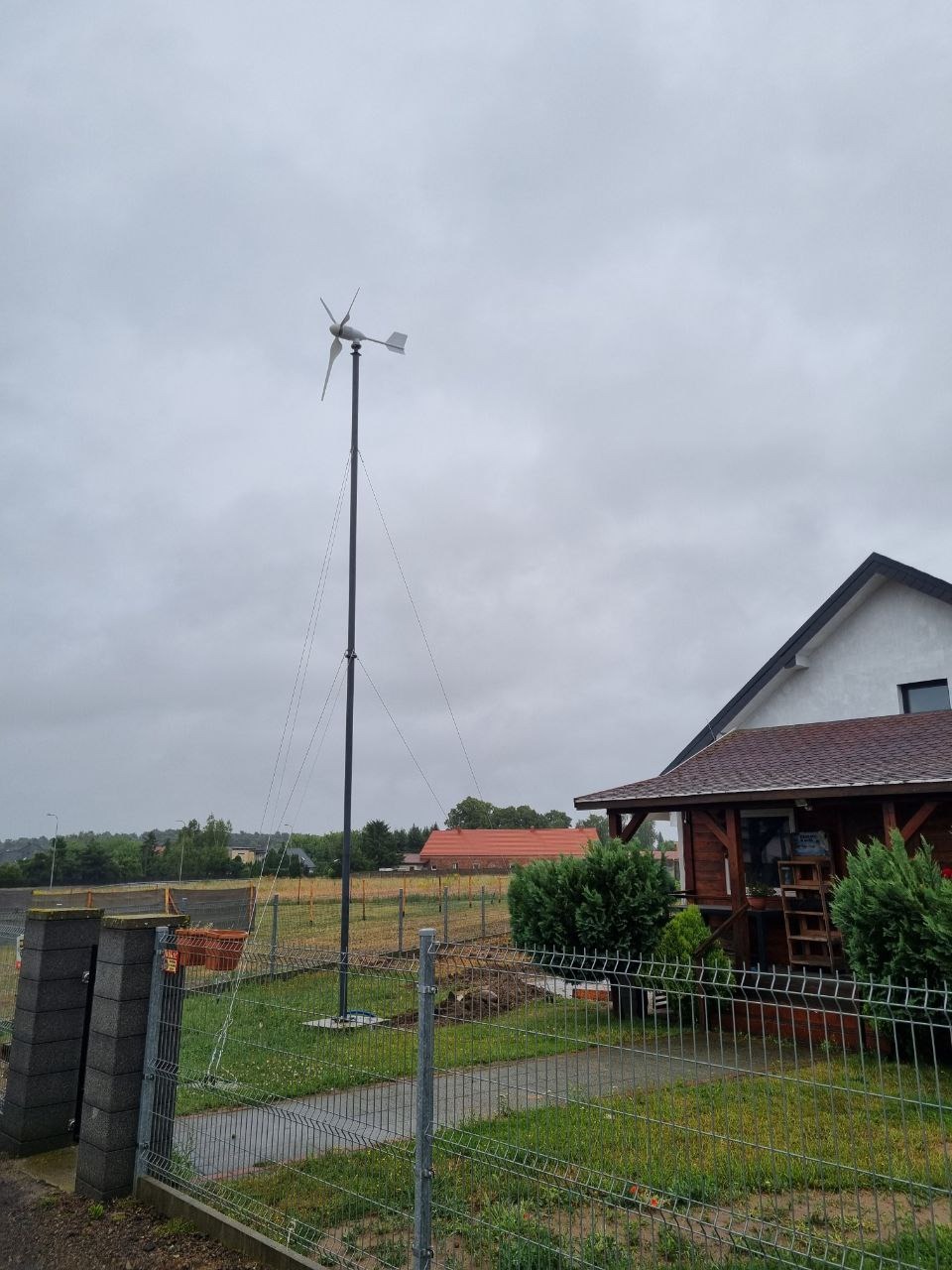 Mała turbina wiatrowa na posesji przy domu jednorodzinnym. Stalowa konstrukcja, trzy łopaty, pochmurne niebo w tle.